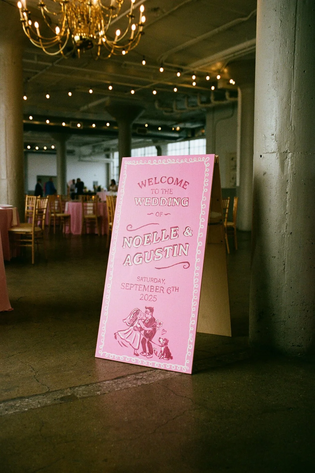 A pink wedding sign with an illustration of a bride and groom dancing and their dog, welcoming guests to Noelle and Agustin's wedding on Saturday, September 6th, 2025, in a rustic indoor venue with round tables, chairs, and string lights.