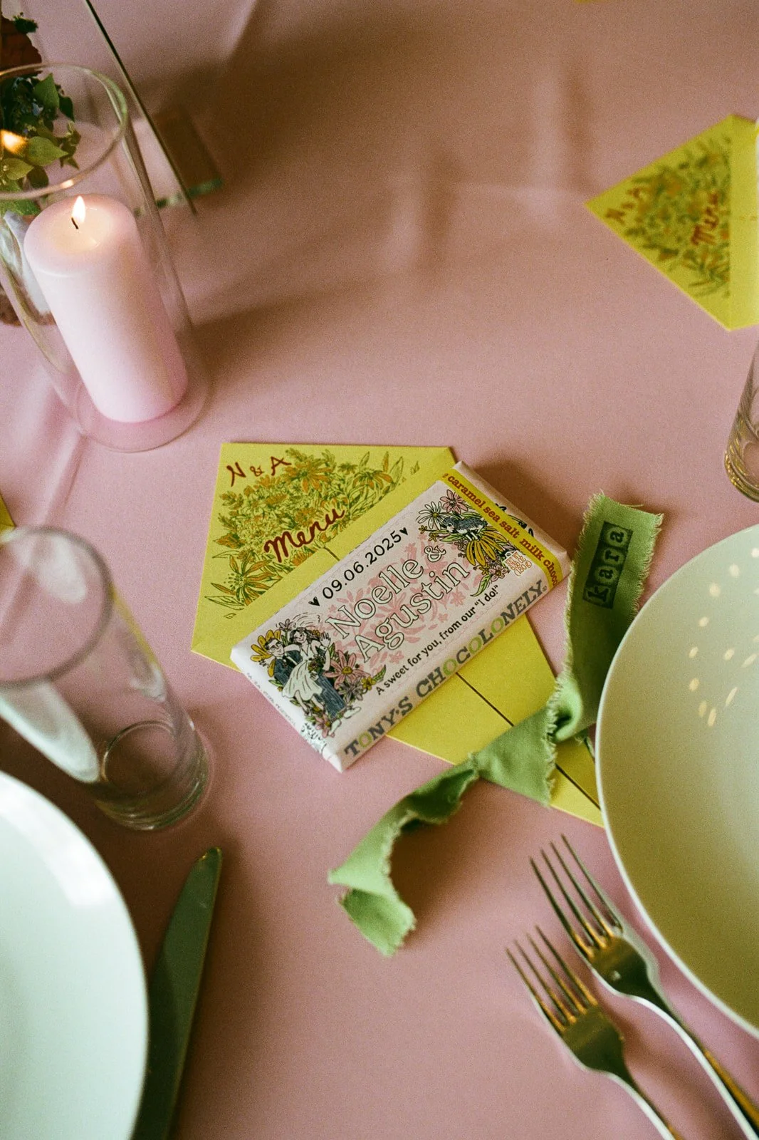 A pink table setting featuring a candle, a menu, a green cloth napkin, and two forks, with some decorative paper and a glass.