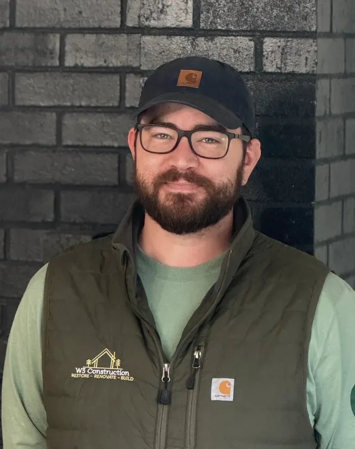 A man with glasses and a beard, wearing a black cap and a green Carhartt vest, standing against a brick wall.