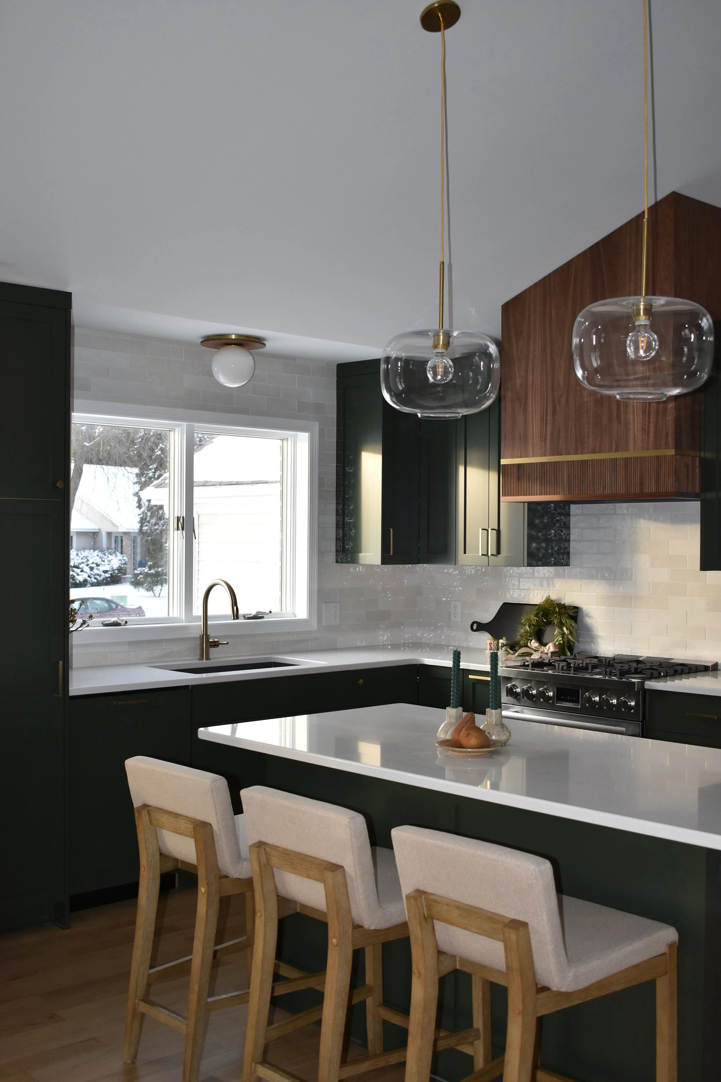 Modern kitchen with dark green cabinets, white countertop, and white backsplash, featuring a window with snowy outdoor scene, two pendant lights with glass globes, and a kitchen island with four beige upholstered chairs.
