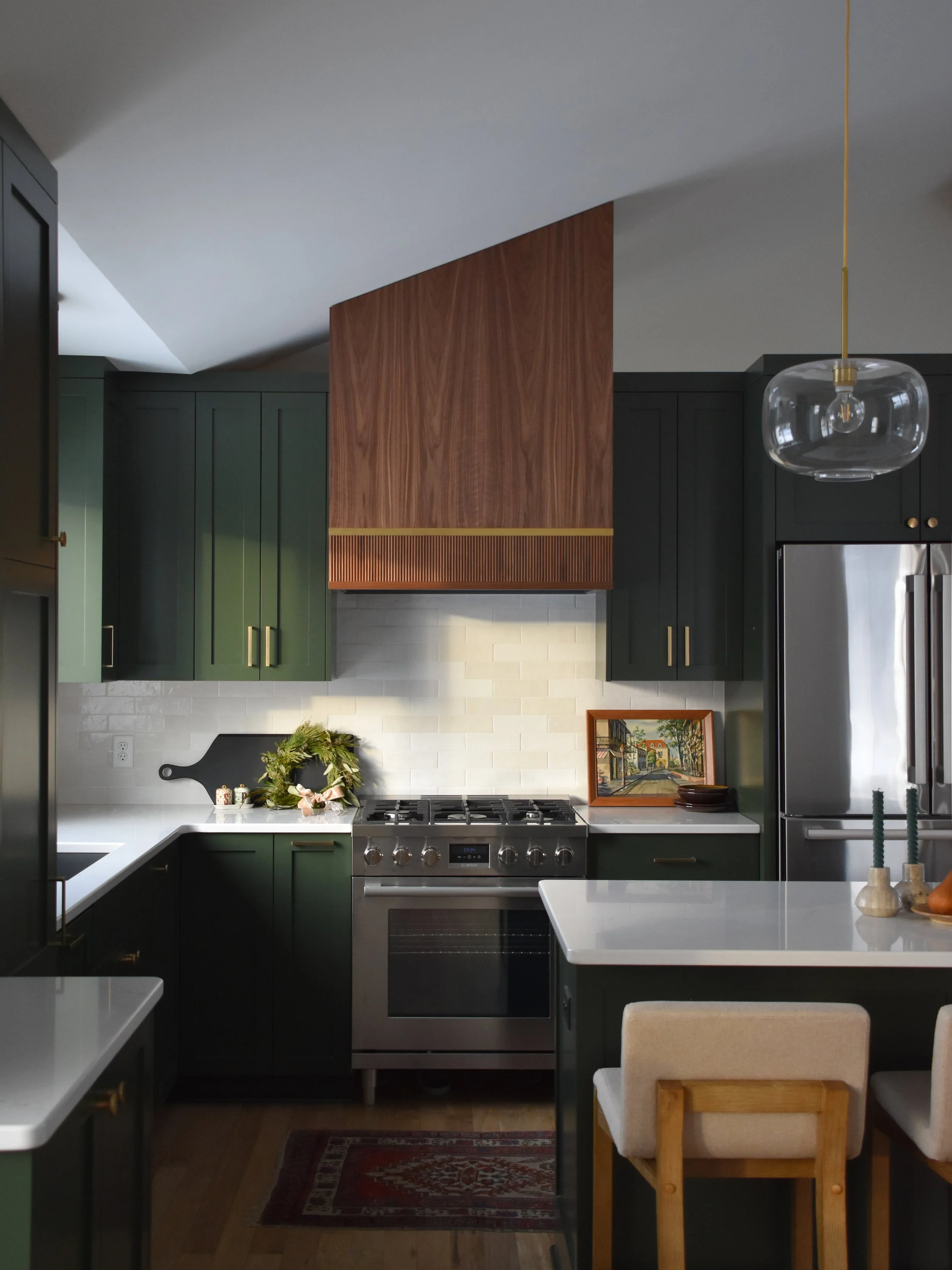 Modern kitchen with dark green cabinets, a white marble countertop, a stainless steel oven, a wooden accent range hood, and a white dining table with beige chairs.