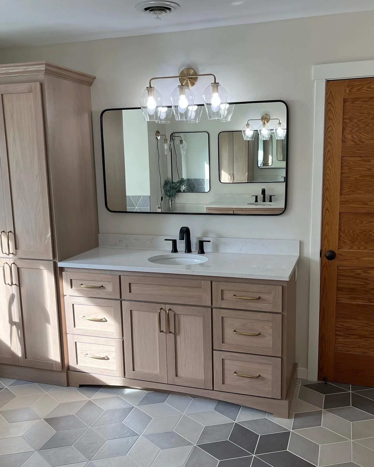 Bathroom vanity with light wooden cabinets, white countertop, black faucet, large mirror, and decorative lighting fixtures mounted on the wall.