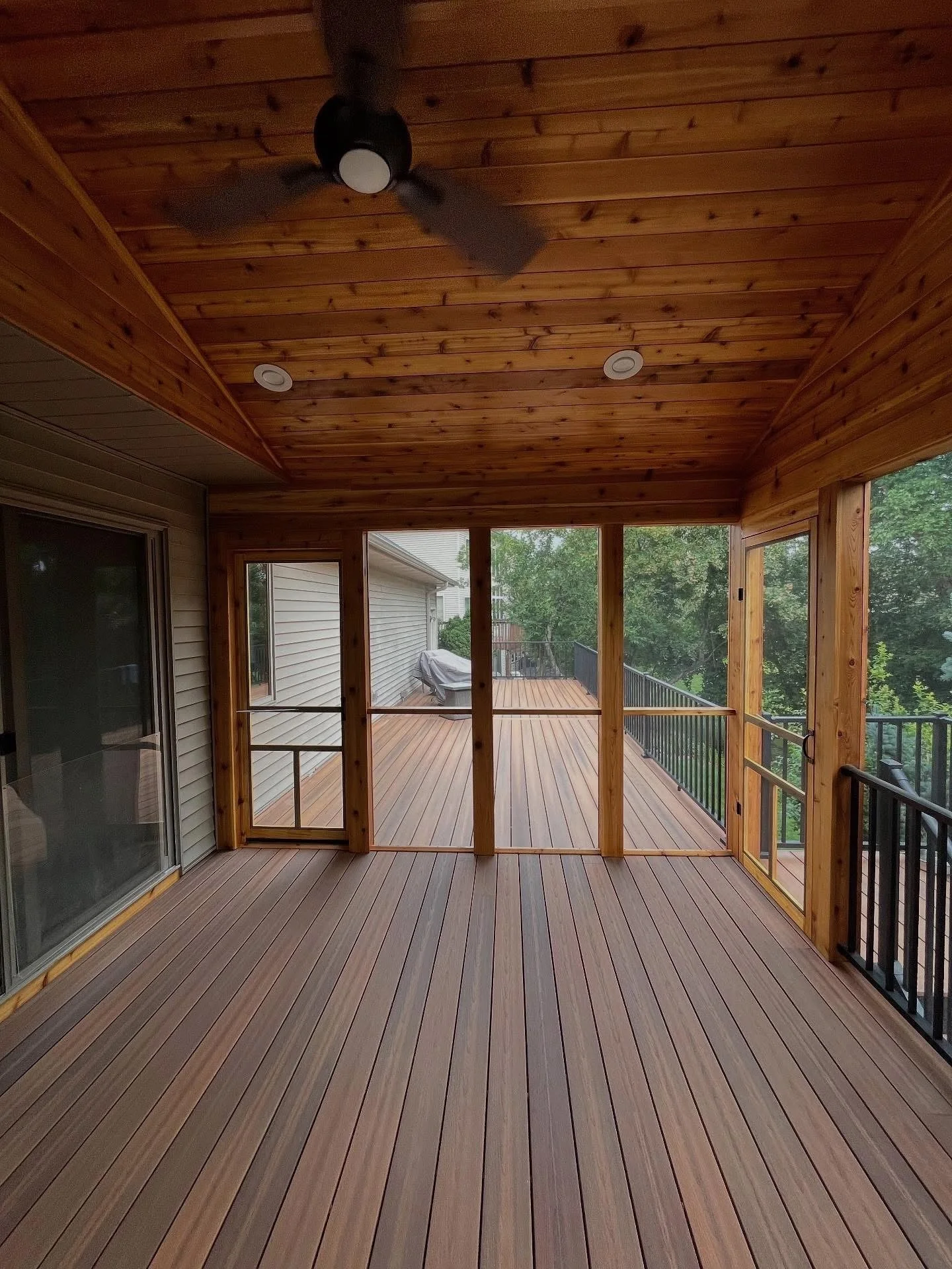 Wooden balcony with sliding glass doors, ceiling fan, and railing, overlooking trees.
