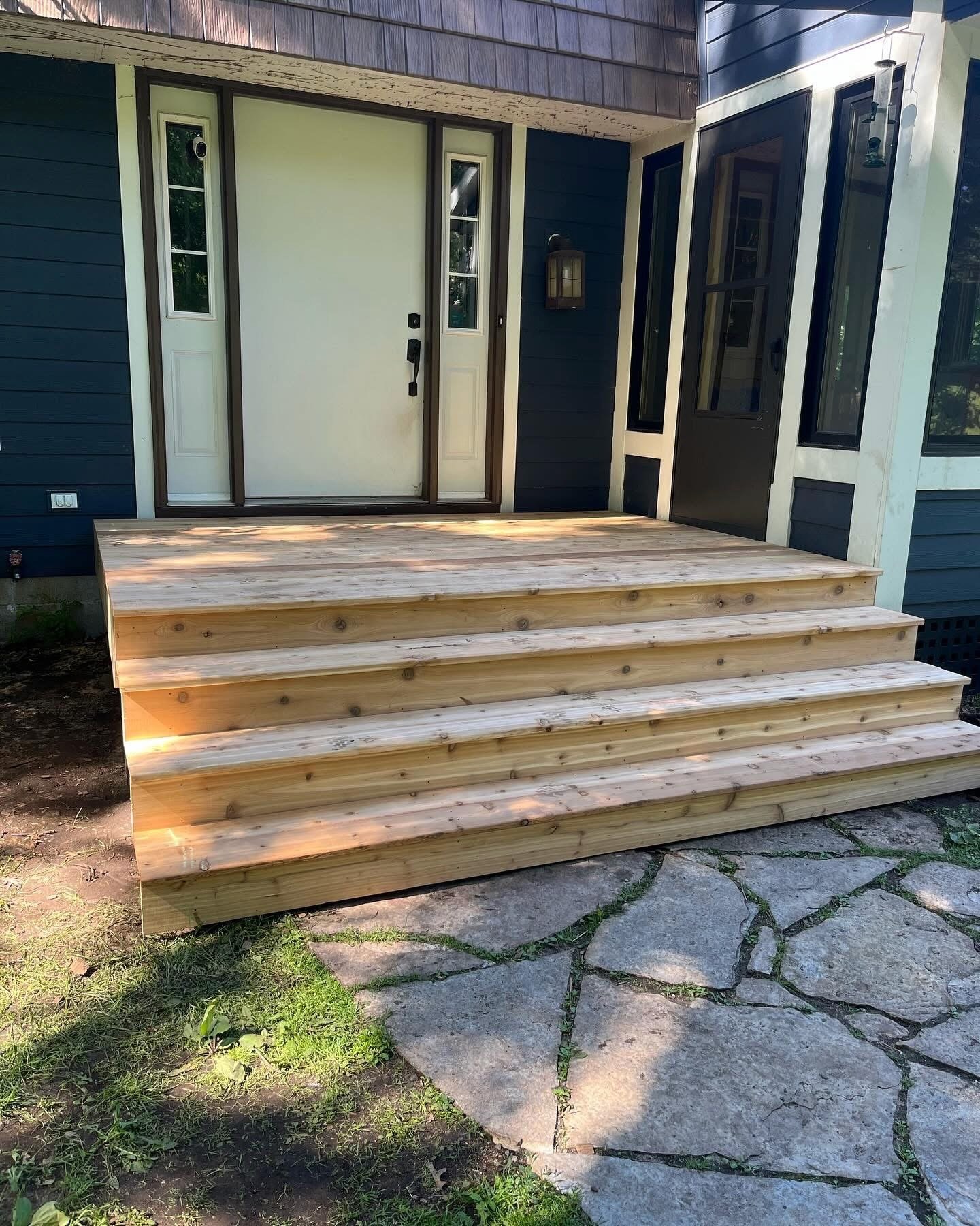 Newly built wooden stairs leading to a house entrance with a door and windows, surrounded by grass and stone pavement.