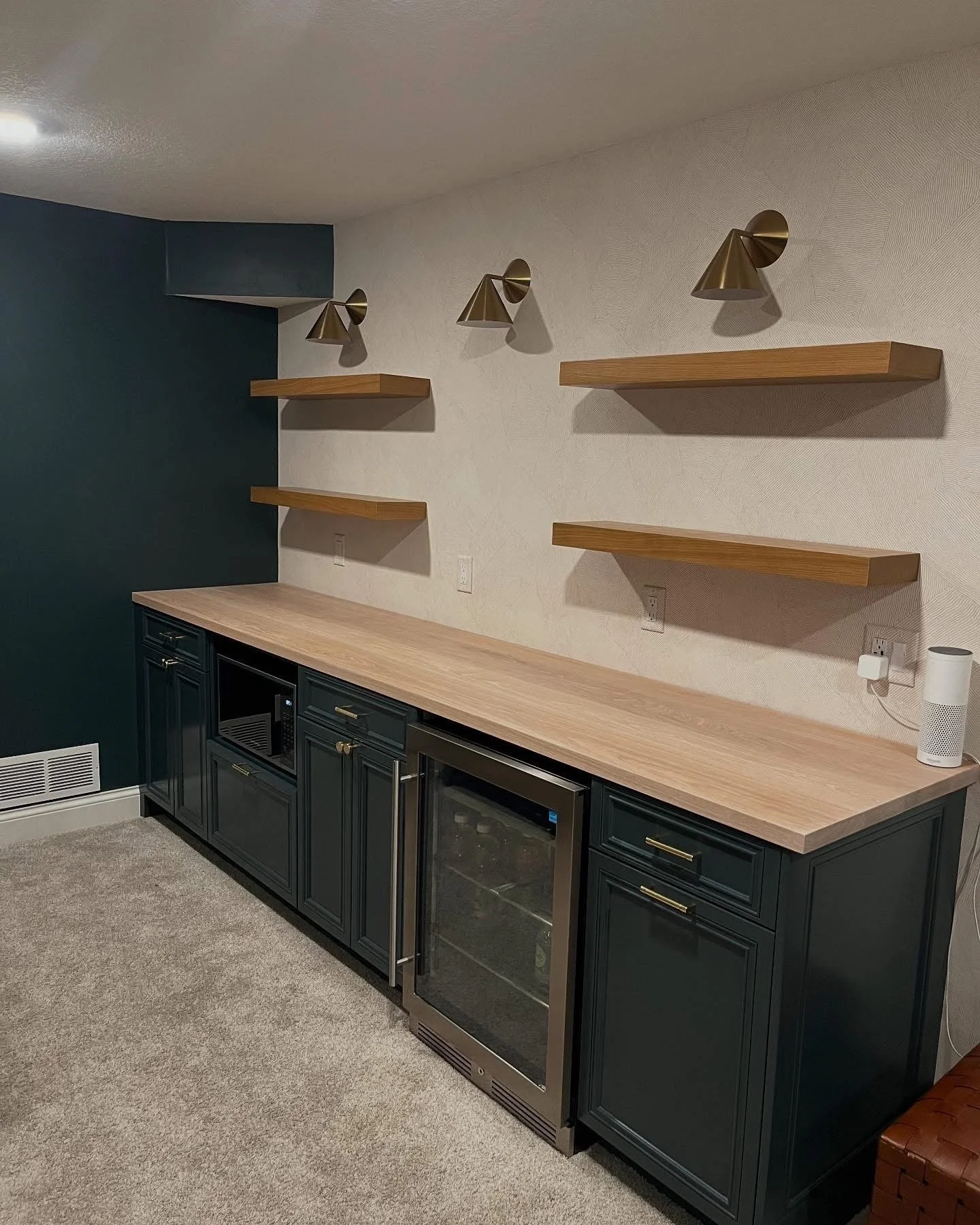 A bar area with dark blue cabinets, a light wood countertop, and a built-in wine or beverage fridge. Three wooden floating shelves are mounted on a white textured wall, with three gold-colored cone-shaped wall sconces above the shelves.