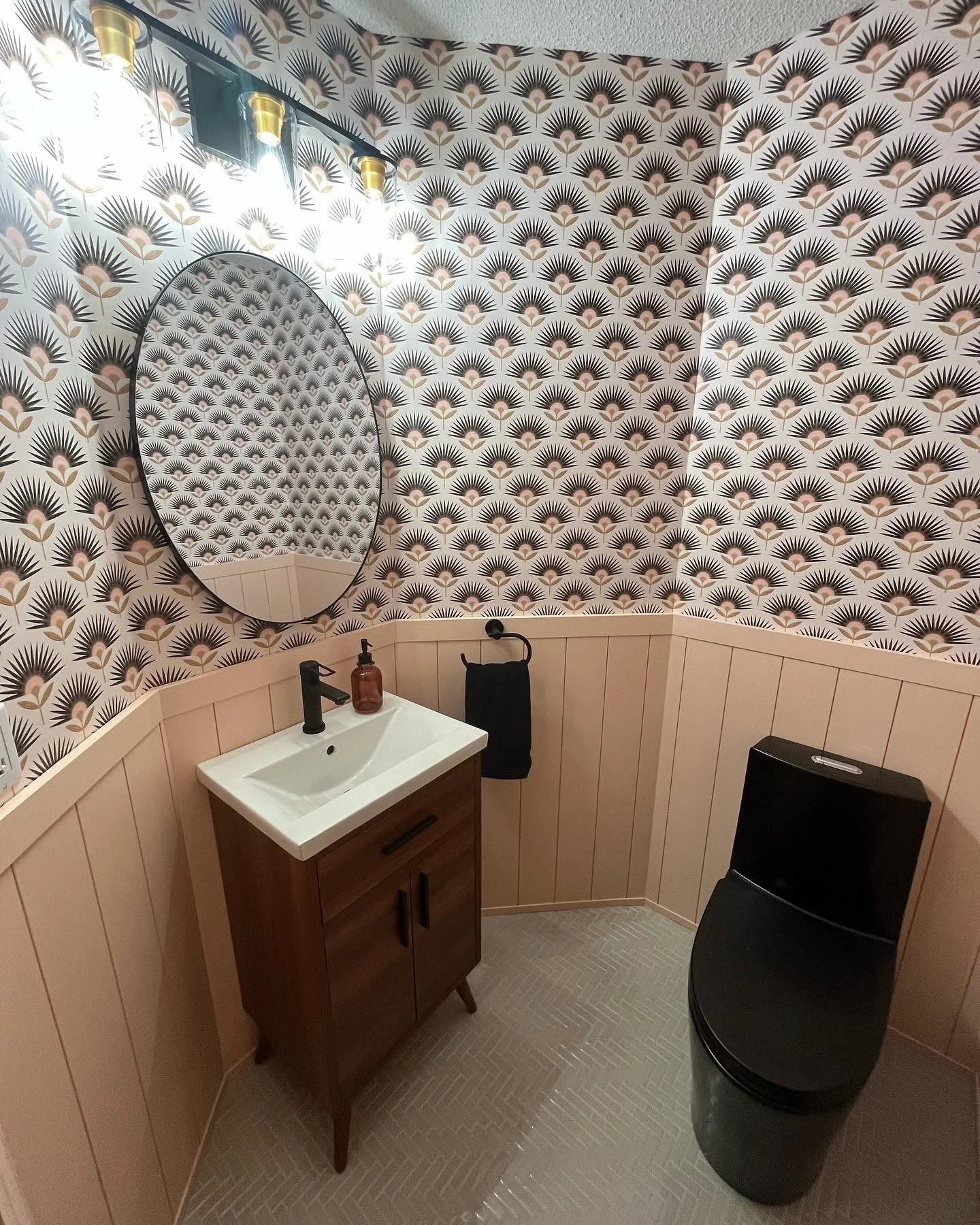 A small bathroom with patterned black and beige wallpaper, a round mirror above a wooden vanity with a white sink, black faucet, a soap dispenser, a black towel on a hook, and a black trash can and a small black appliance in the corner, with light-co