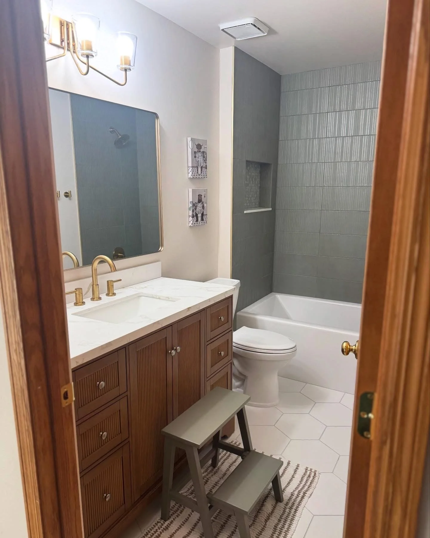 A bathroom with a wooden vanity, marble countertop, gold fixtures, a large mirror, wall art, a toilet, and a bathtub with gray tile walls.