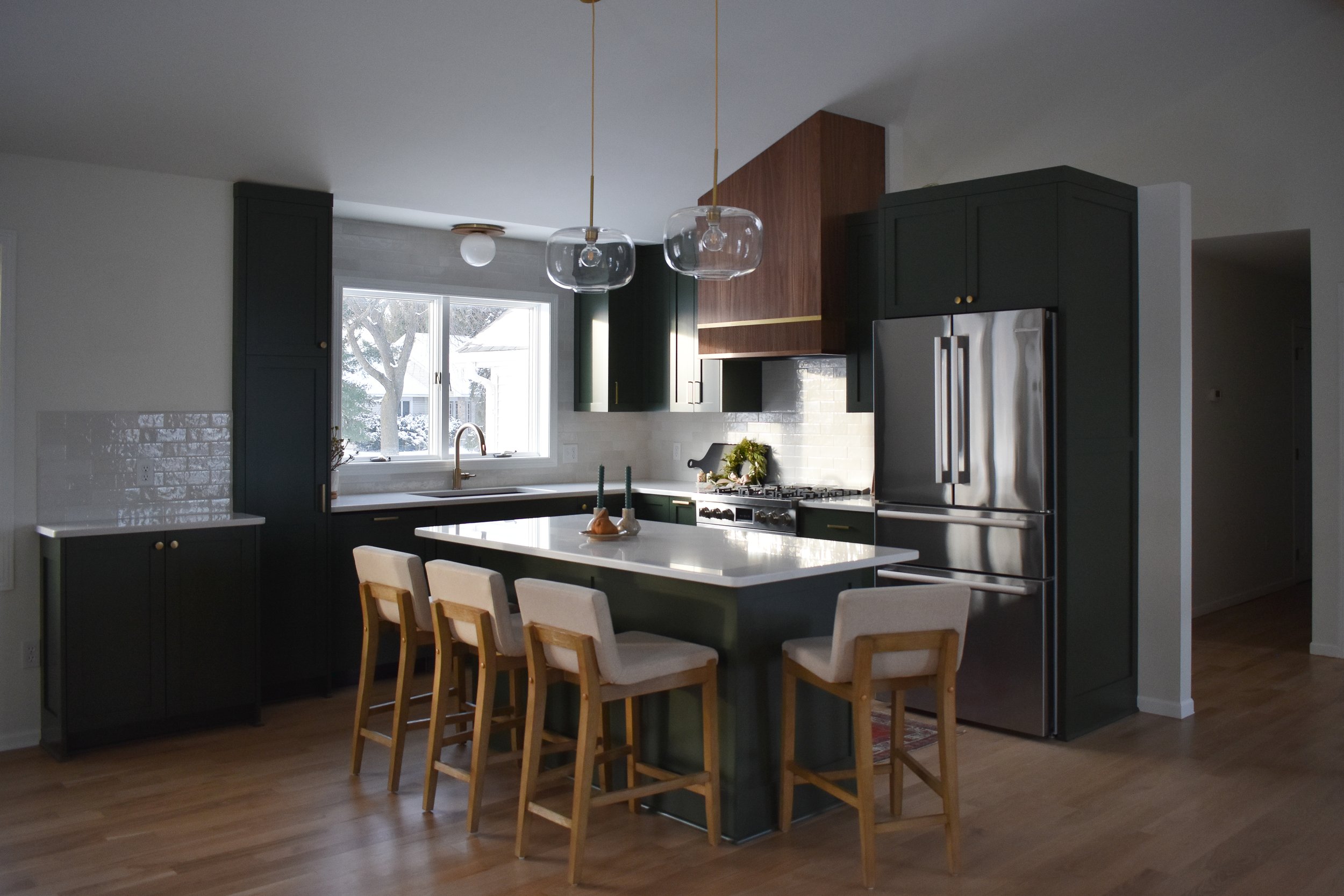 Modern kitchen with dark green cabinets, white countertops, a stainless steel refrigerator, a gas stove, and a large window with trees outside. There are five chairs around a kitchen island with candles and a decorative tray, pendant lights hang from the ceiling, and the floor is wood.