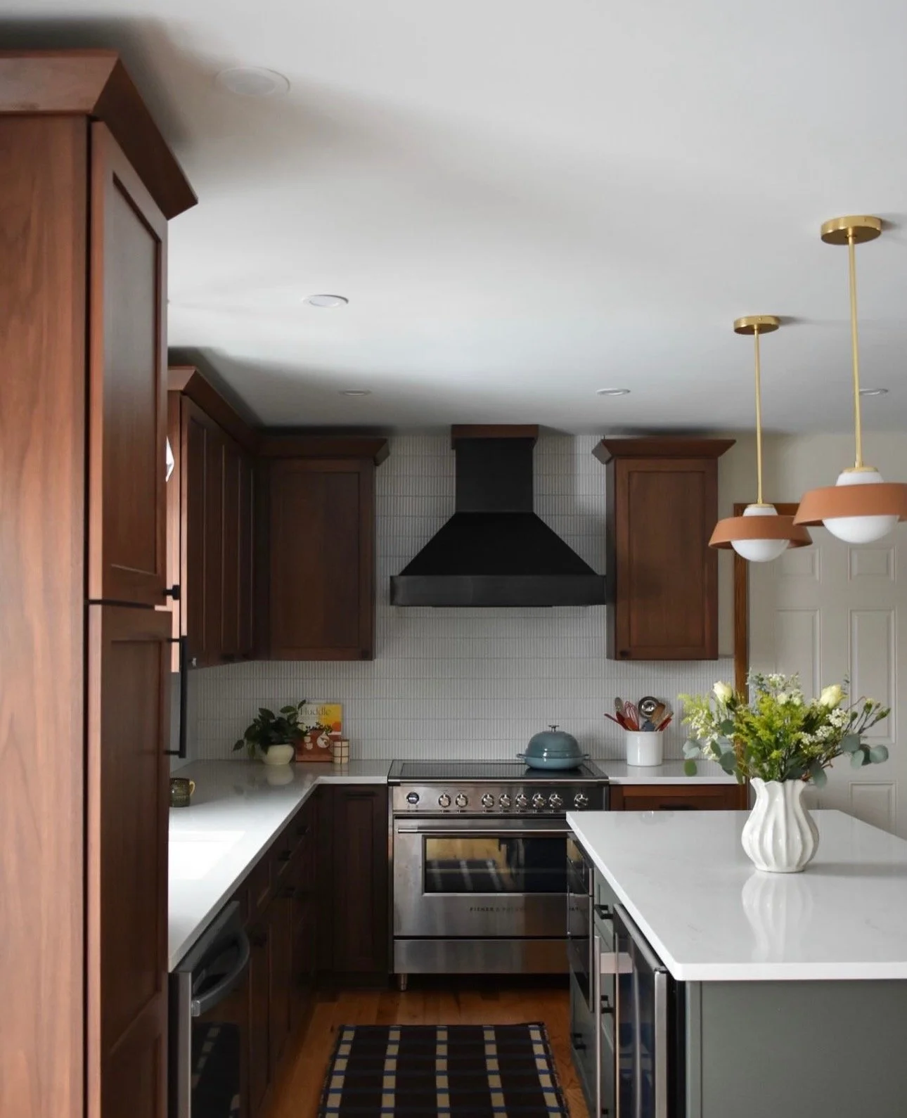 A modern kitchen with dark wood cabinets, stainless steel oven, white countertops, a black range hood, and a white tiled backsplash. There is a white vase with flowers, pendant lights, and a few decorative items on the counter.