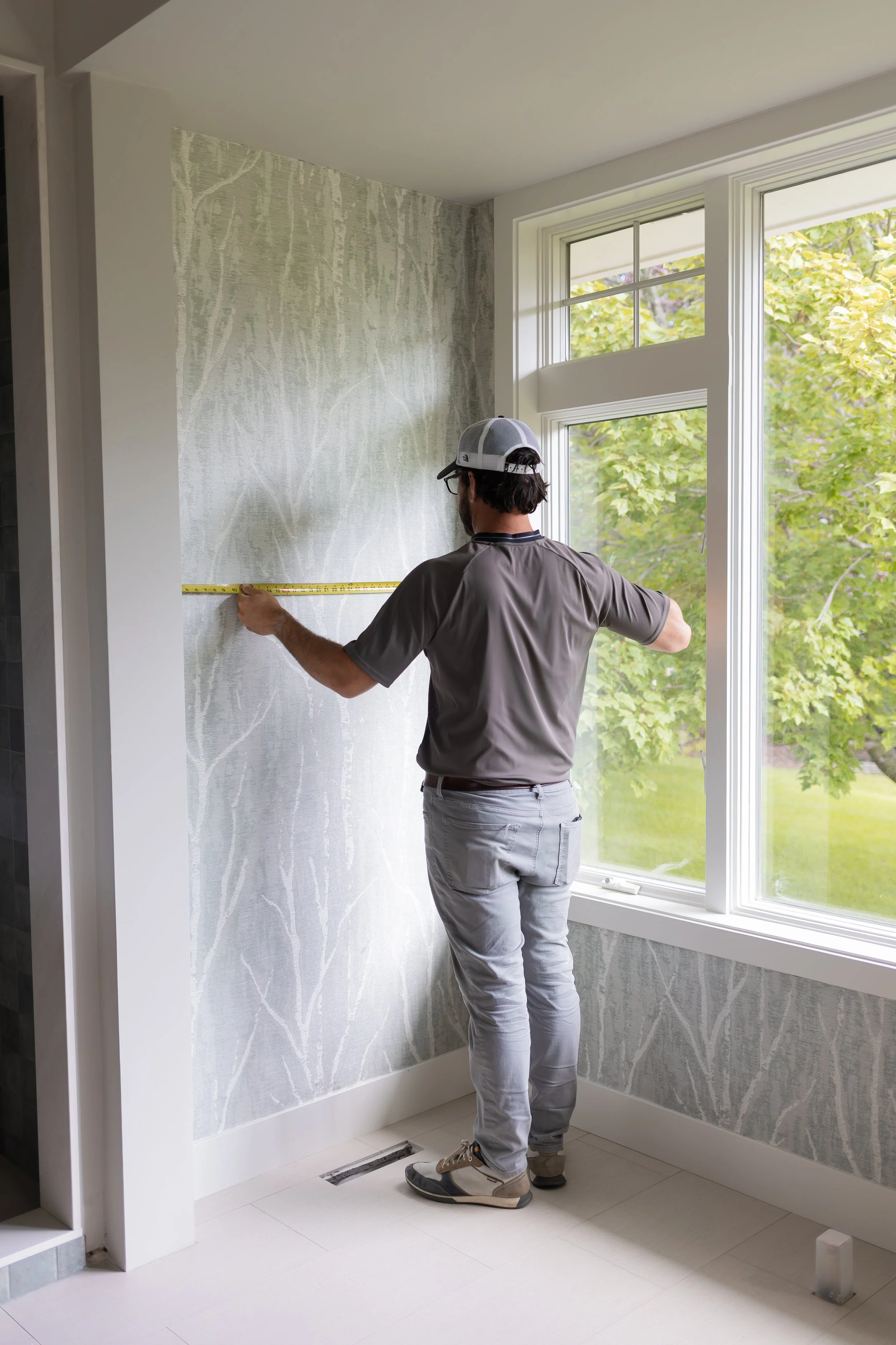 A man measuring a wall with a tape measure in a room with large bay windows and patterned wallpaper.