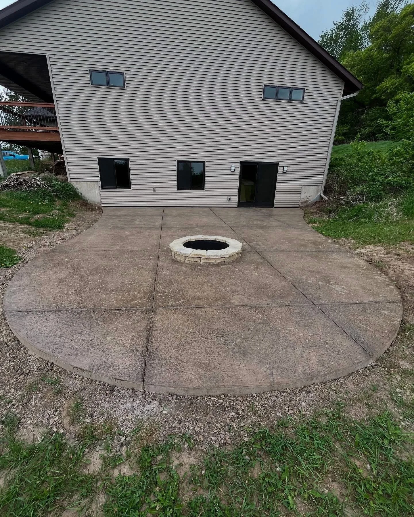 A circular concrete patio with a fire pit in the center, attached to the back of a beige house with a sliding glass door and three small windows.