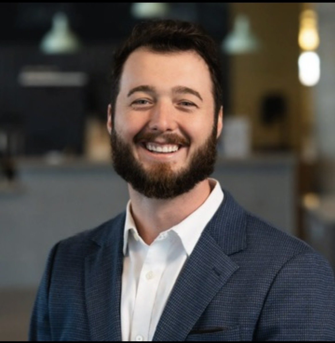 Headshot of a smiling man with a beard, wearing a blue blazer and white shirt, in a professional indoor setting.