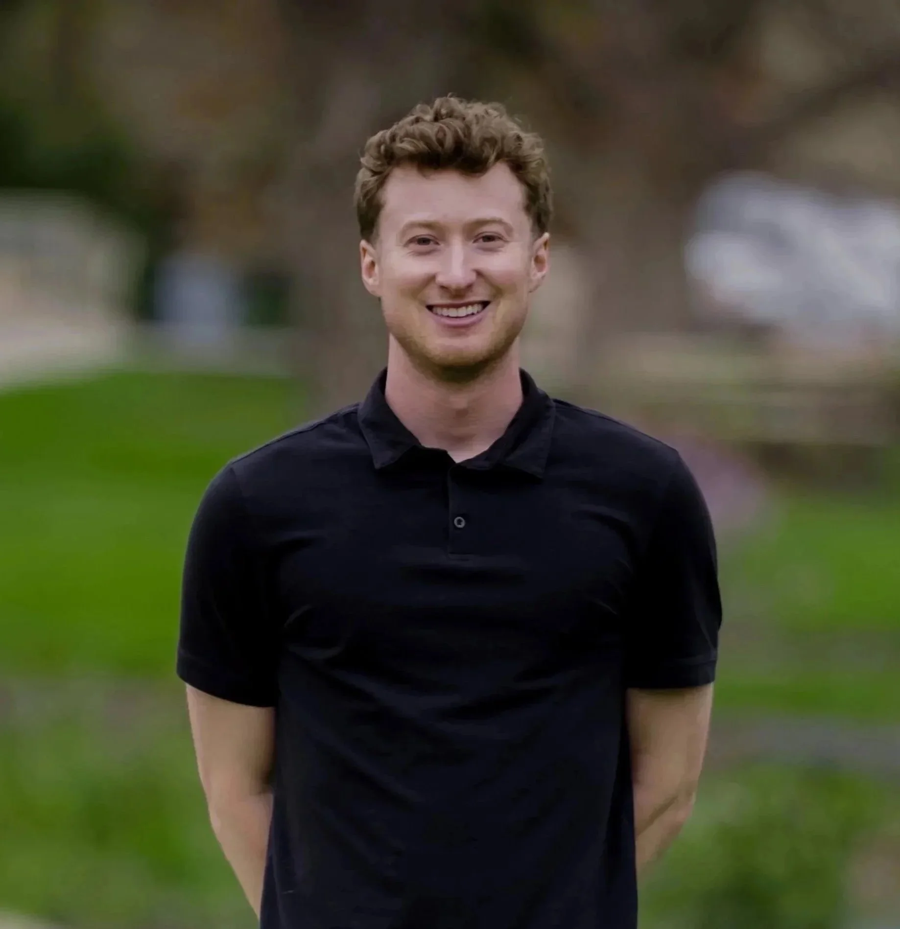 A smiling young man with curly hair wearing a black shirt outdoors on a cloudy day.