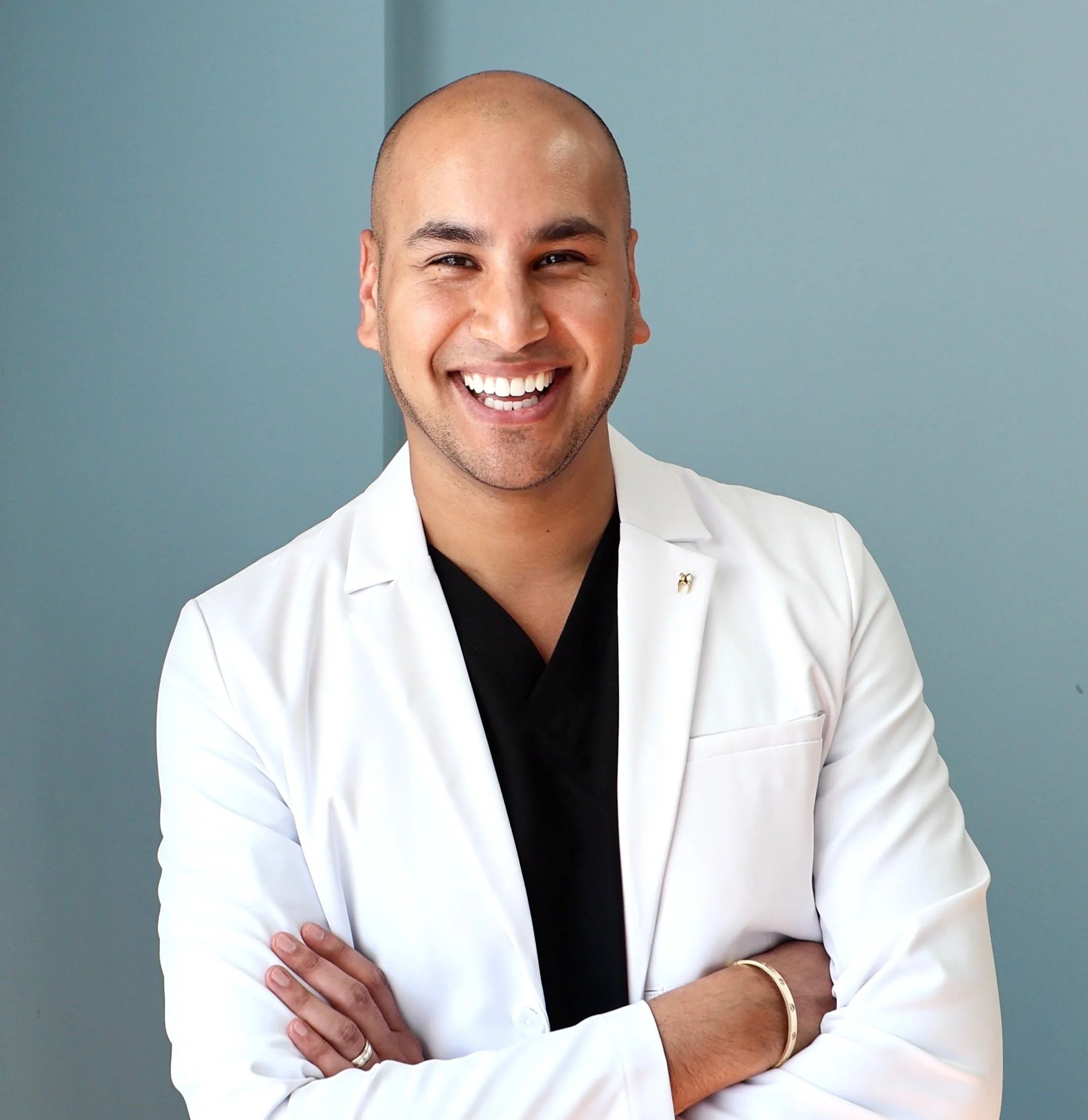Smiling man with crossed arms wearing a white coat, standing in front of a light blue wall.