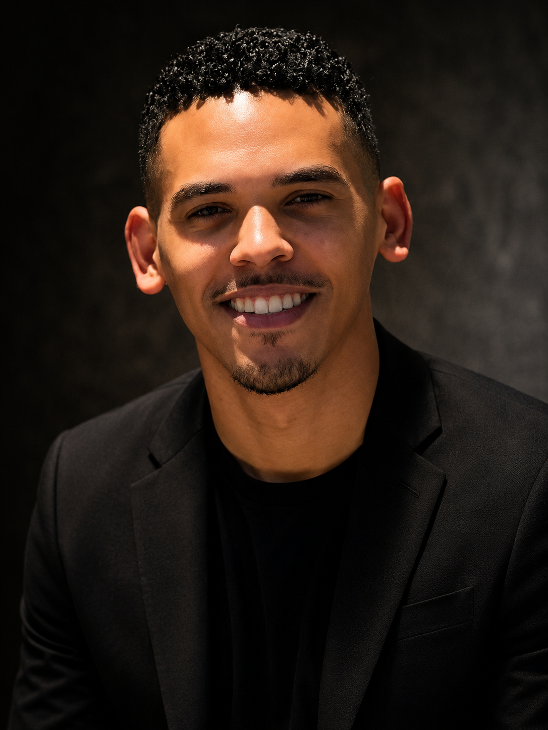 Young man with short curly black hair, light skin, and a trimmed goatee, smiling in a dark setting, wearing a black blazer over a black shirt.