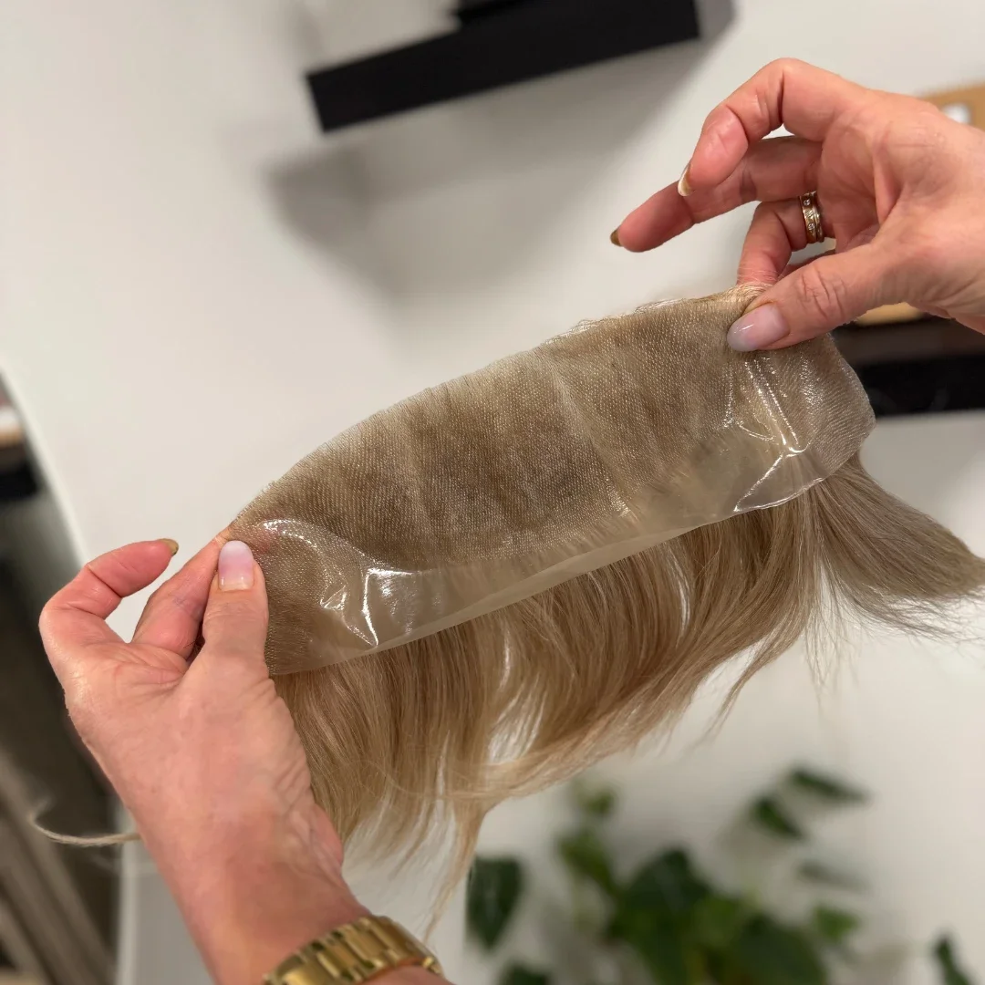 Hands holding a blonde hair wig with a lace front, partially covered by a transparent adhesive lace wig cap.