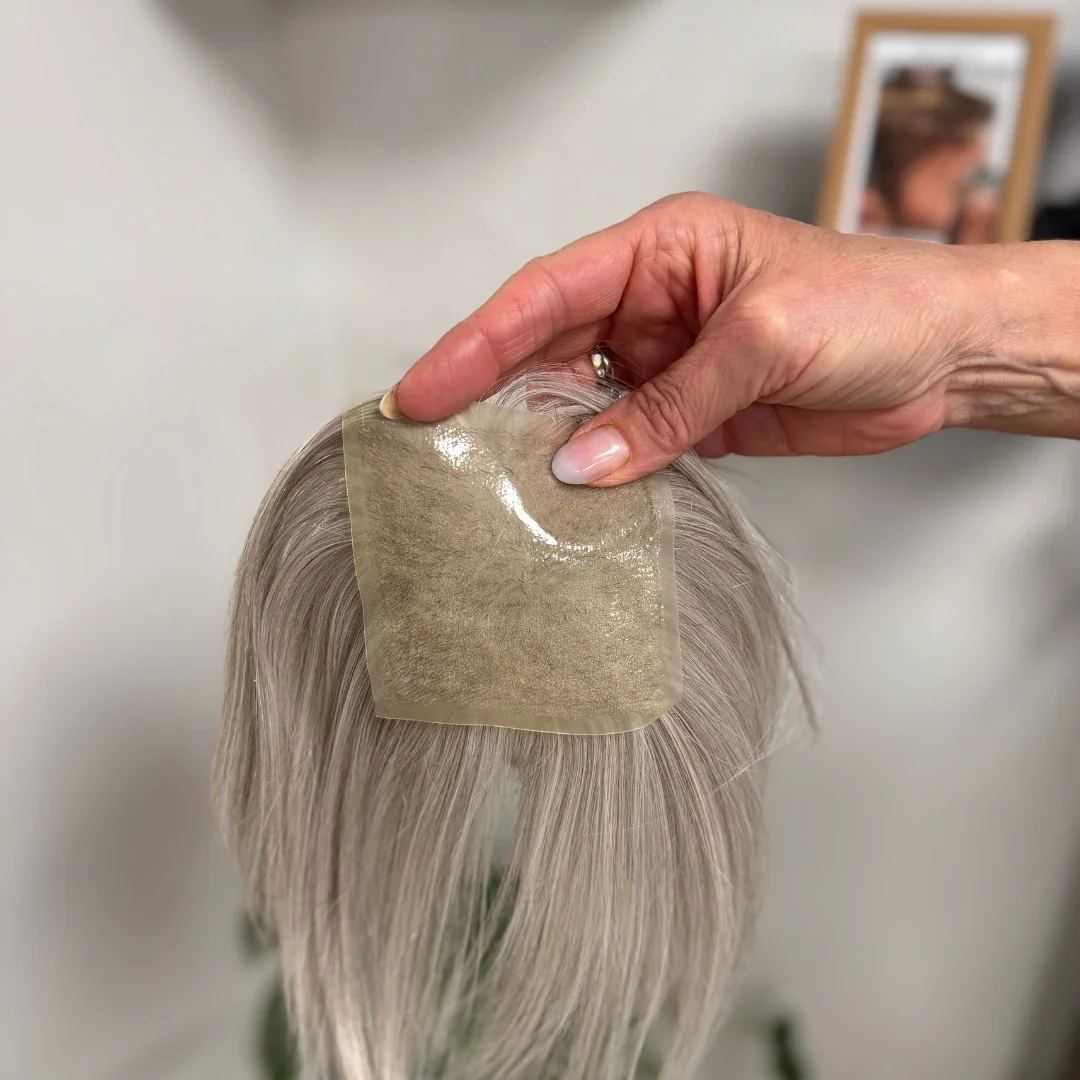 A person is applying a hair extension patch to the top of a person's head with light blonde hair.