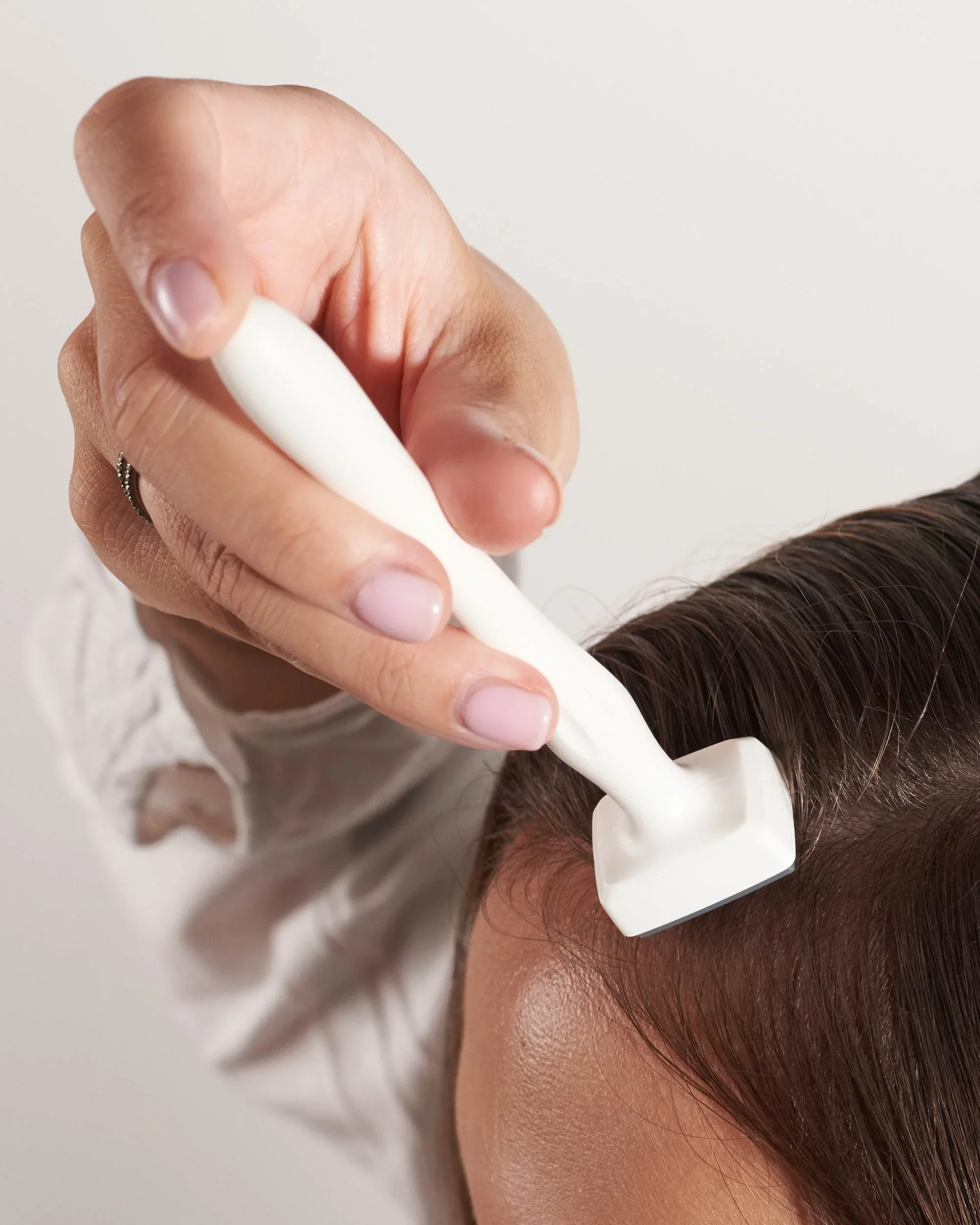 A person receiving a scalp treatment with a white handheld device on their dark brown hair and scalp, in a close-up view.