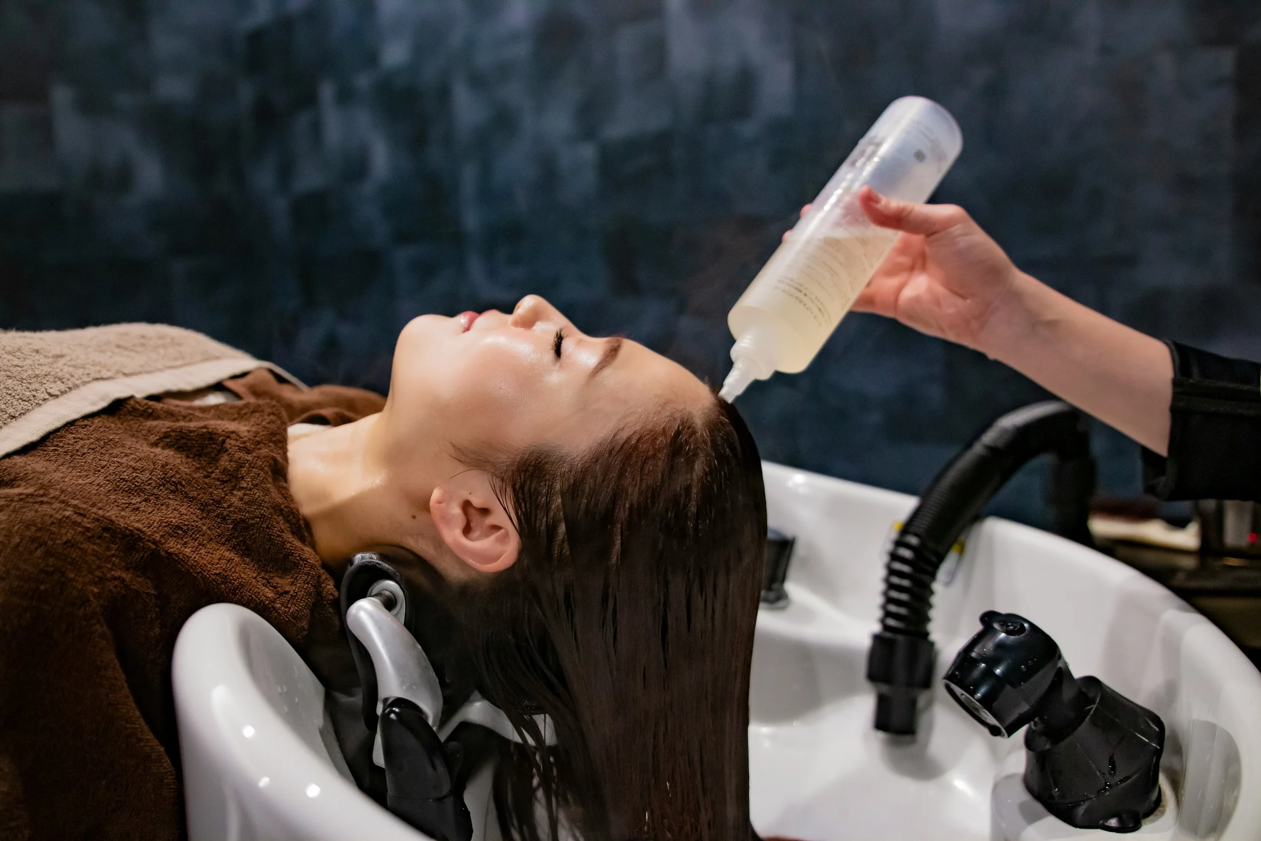 A woman lying back in a salon chair while a stylist applies shampoo or conditioner to her hair from a clear bottle.