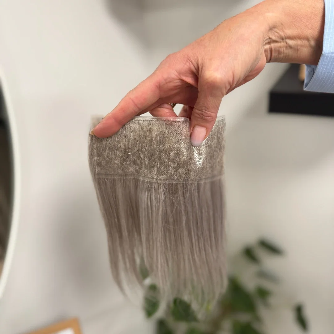 A hand holding a sample of beige hair extension or wig with a transparent top and silky strands.