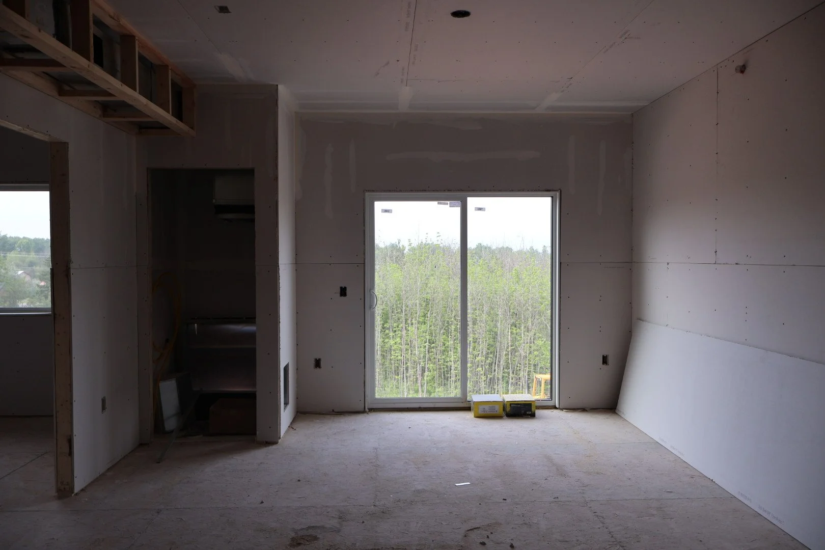 Interior of a house under construction with unfinished drywall, open wall frames, a large window showing a treed landscape, and construction materials on the floor.