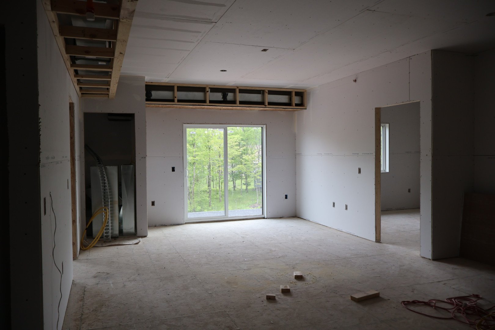 Photo of an unfinished room under construction, with drywall on the walls, a sliding glass door leading outside, and a small window. Construction materials and wires are visible, and the ceiling has exposed framing for lighting fixtures.