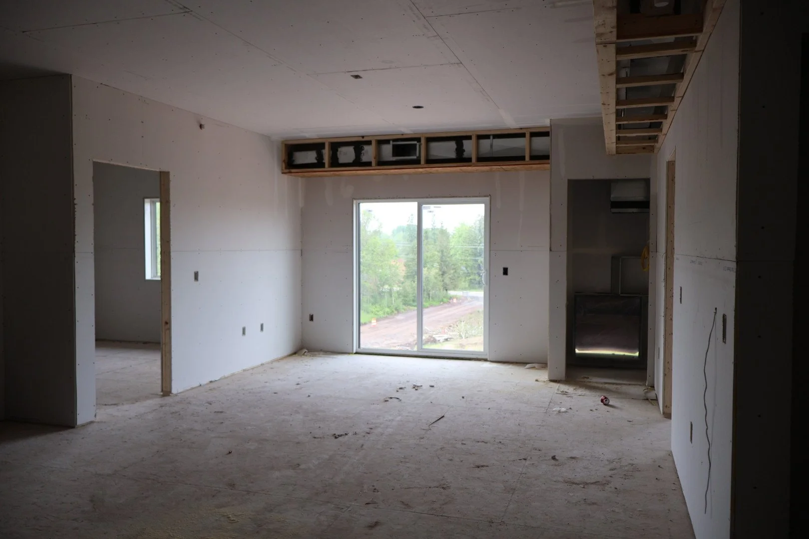 Interior view of a house under construction with unfinished drywall, exposed ceiling and wall framing, sliding glass door leading outside, and construction debris on the floor.