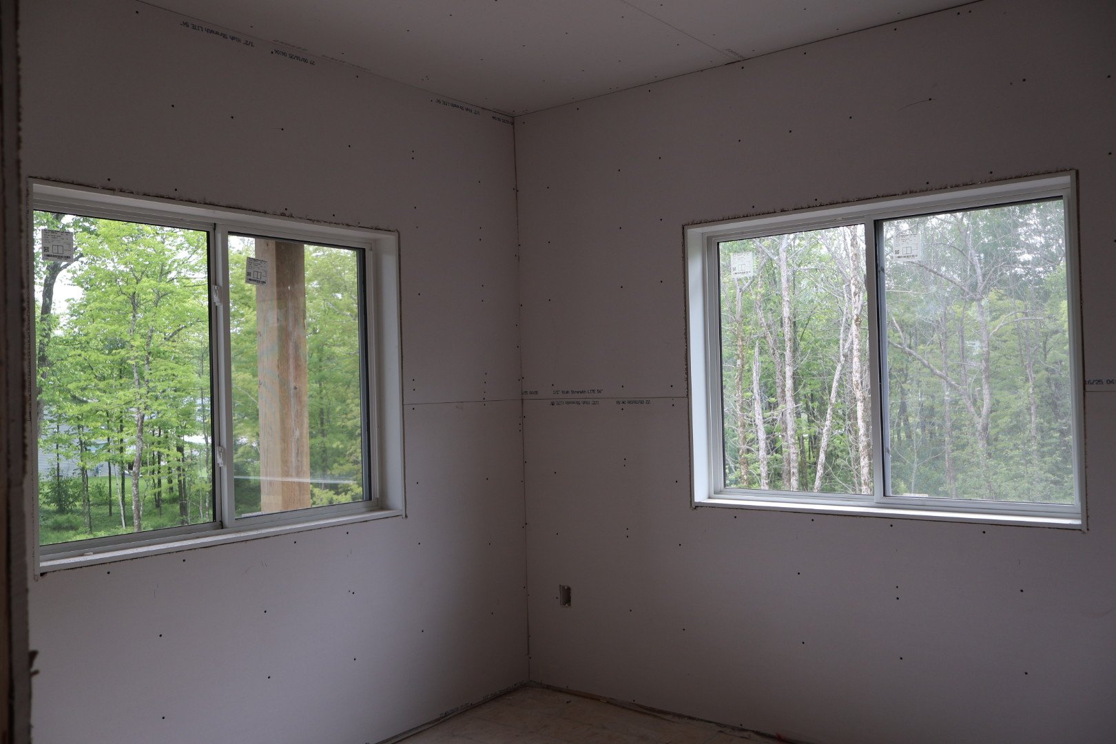Interior of an unfinished room with two large windows showing green trees outside.