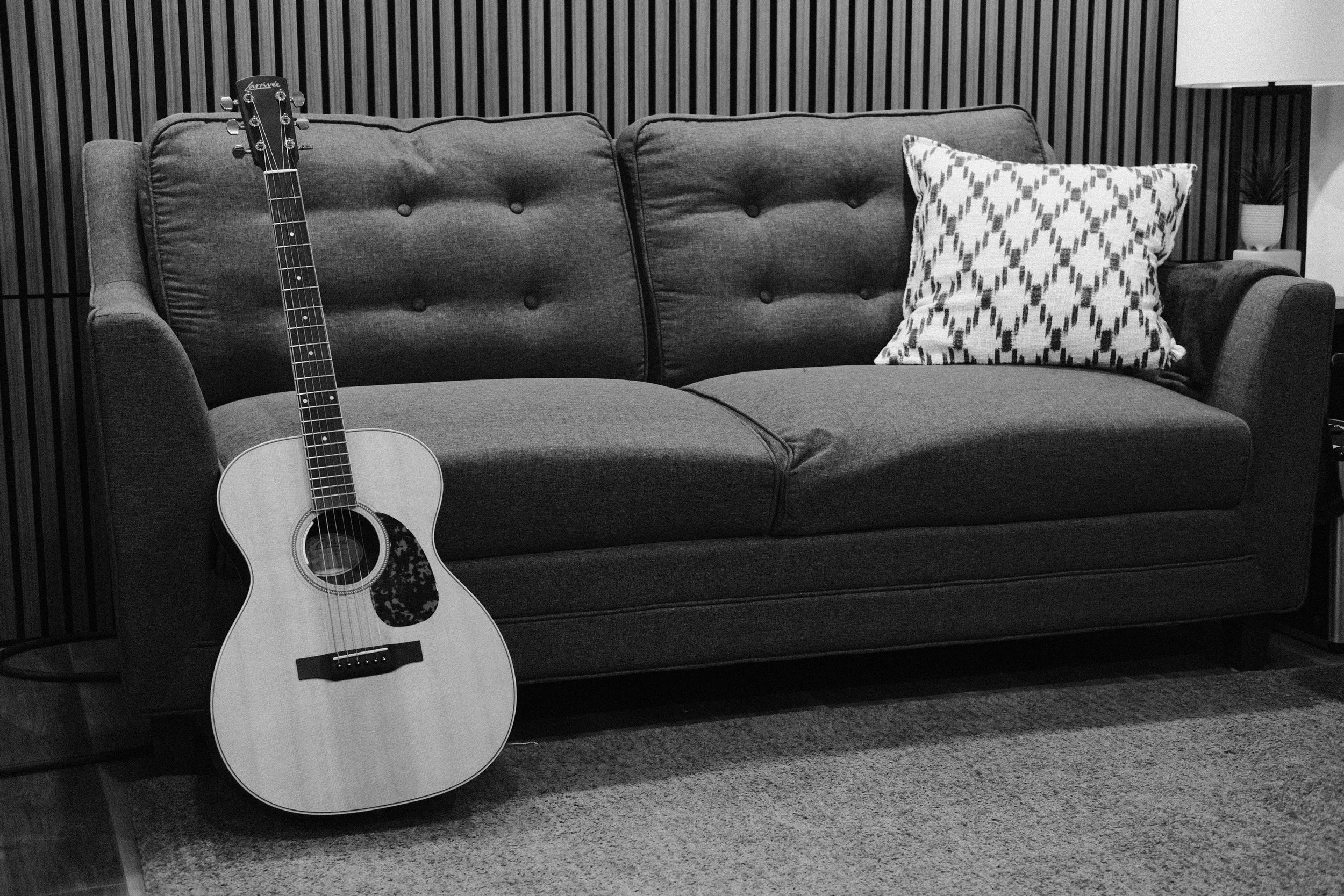 A grayscale photo of a living room with a dark fabric sofa, a patterned pillow, a wooden acoustic guitar leaning against the left arm, a lamp, and a potted plant in the background.