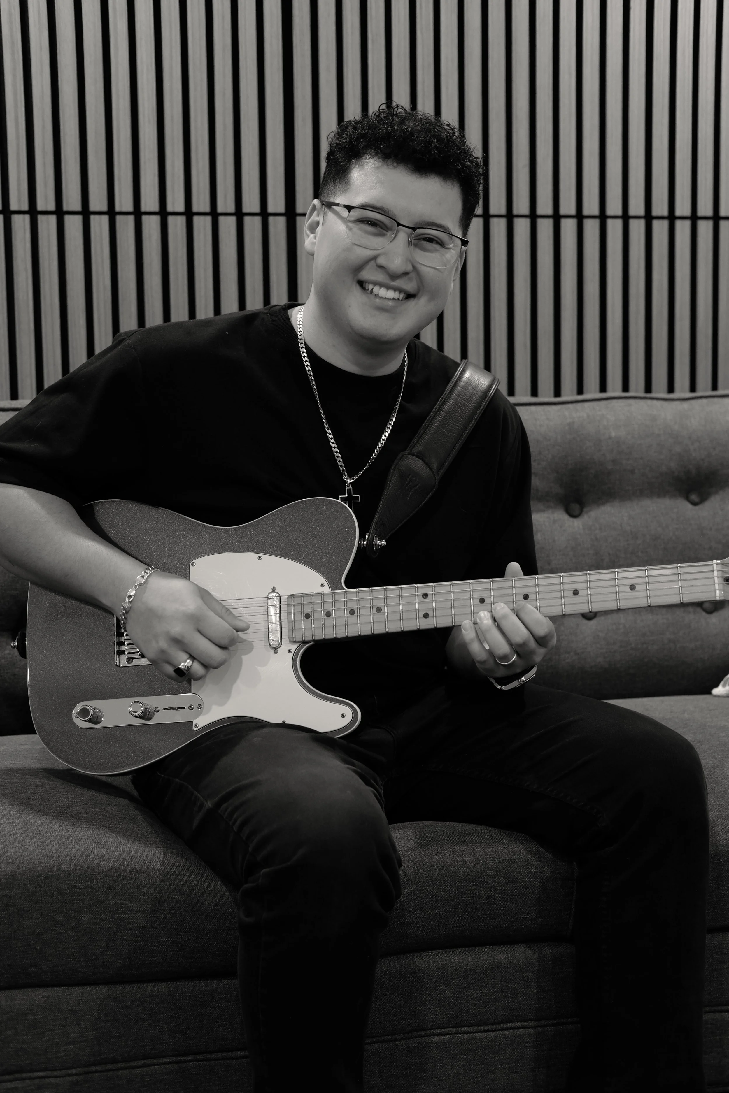 A young man sitting on a couch, smiling, holding an electric guitar, wearing glasses, a chain with a cross, and a bracelet, with a wooden slat wall background.