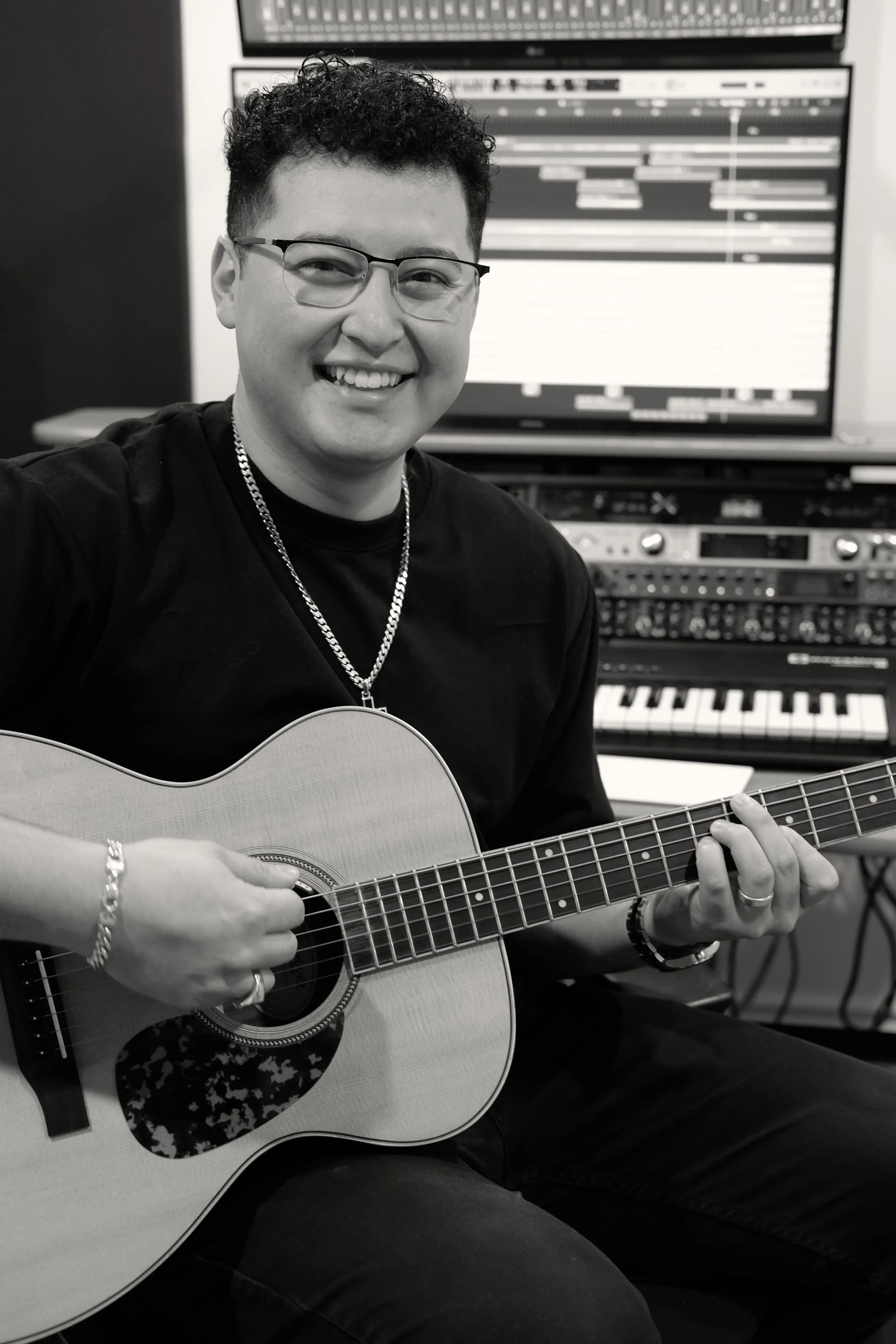A man with glasses and earrings smiling while playing an acoustic guitar in a music studio, with music production equipment and a computer monitor with digital audio workstations in the background.