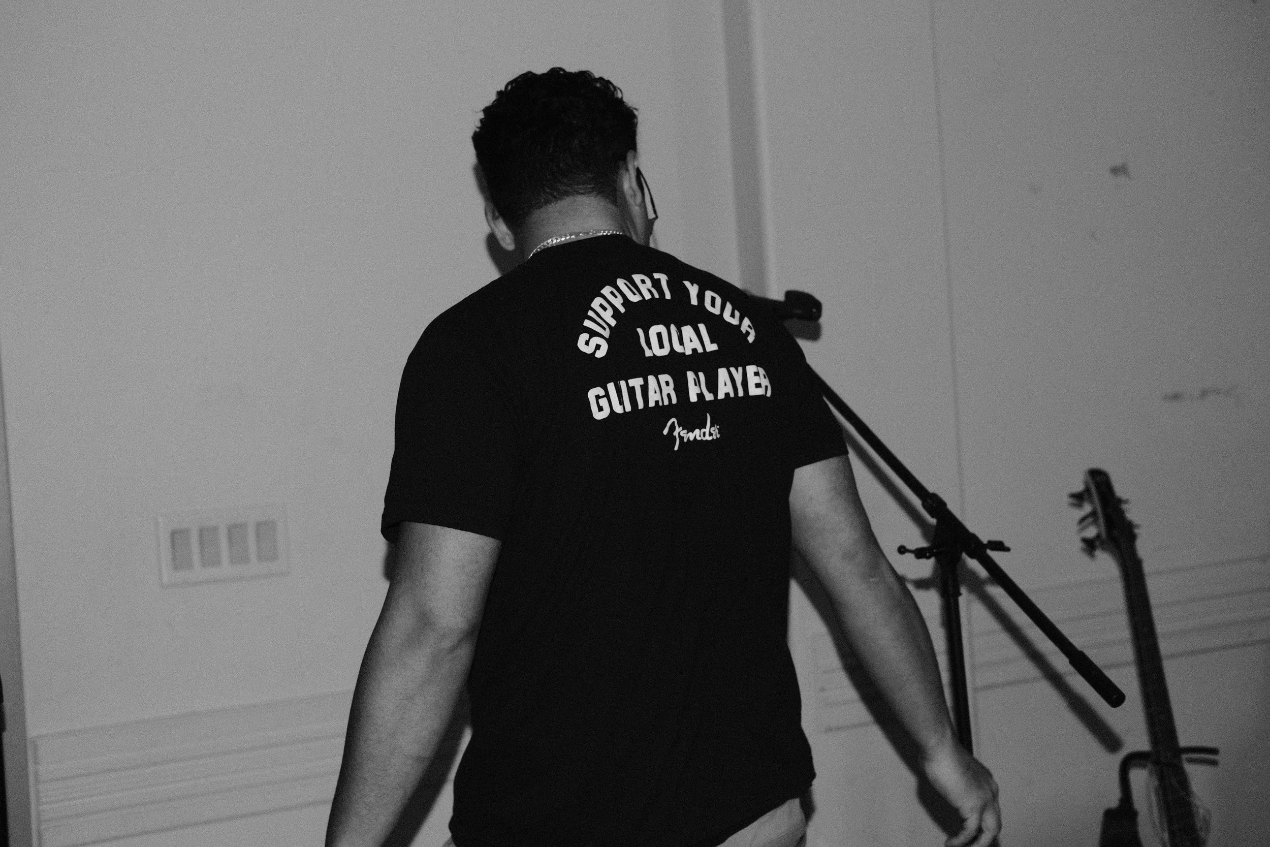 Back view of a young man with short dark hair, wearing a black T-shirt with white text, supporting a guitar player, indoors with a plain wall and a guitar stand in the background.