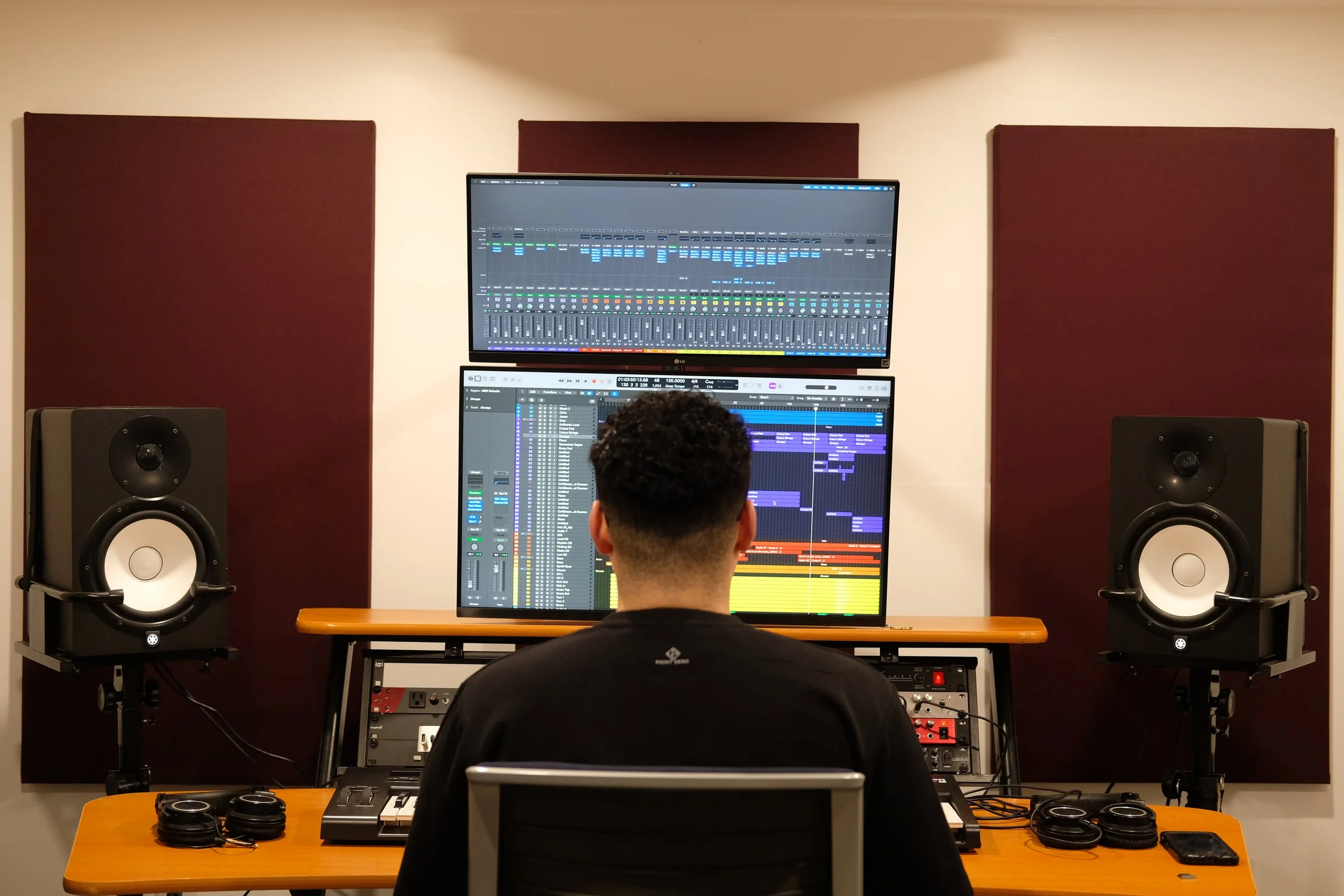 Person sitting at a desk in a music studio, working on a digital audio workstation on a dual monitor setup, with studio speakers and audio equipment around.