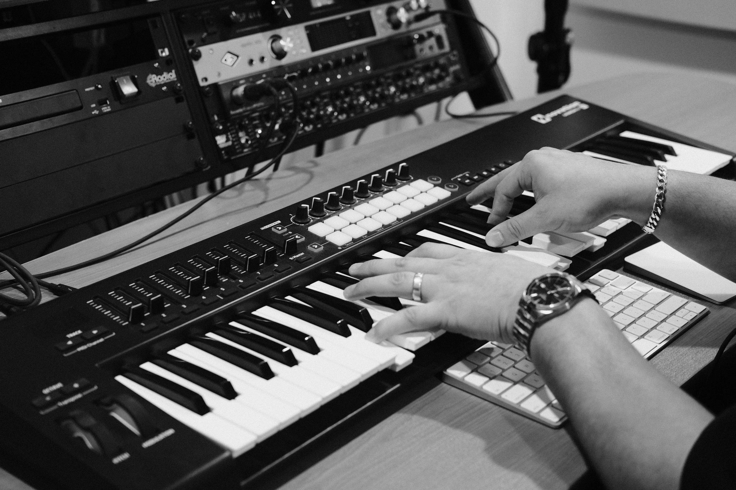 Black and white photo of a person's hands playing a keyboard, surrounded by audio equipment, including a mixer and a computer keyboard.