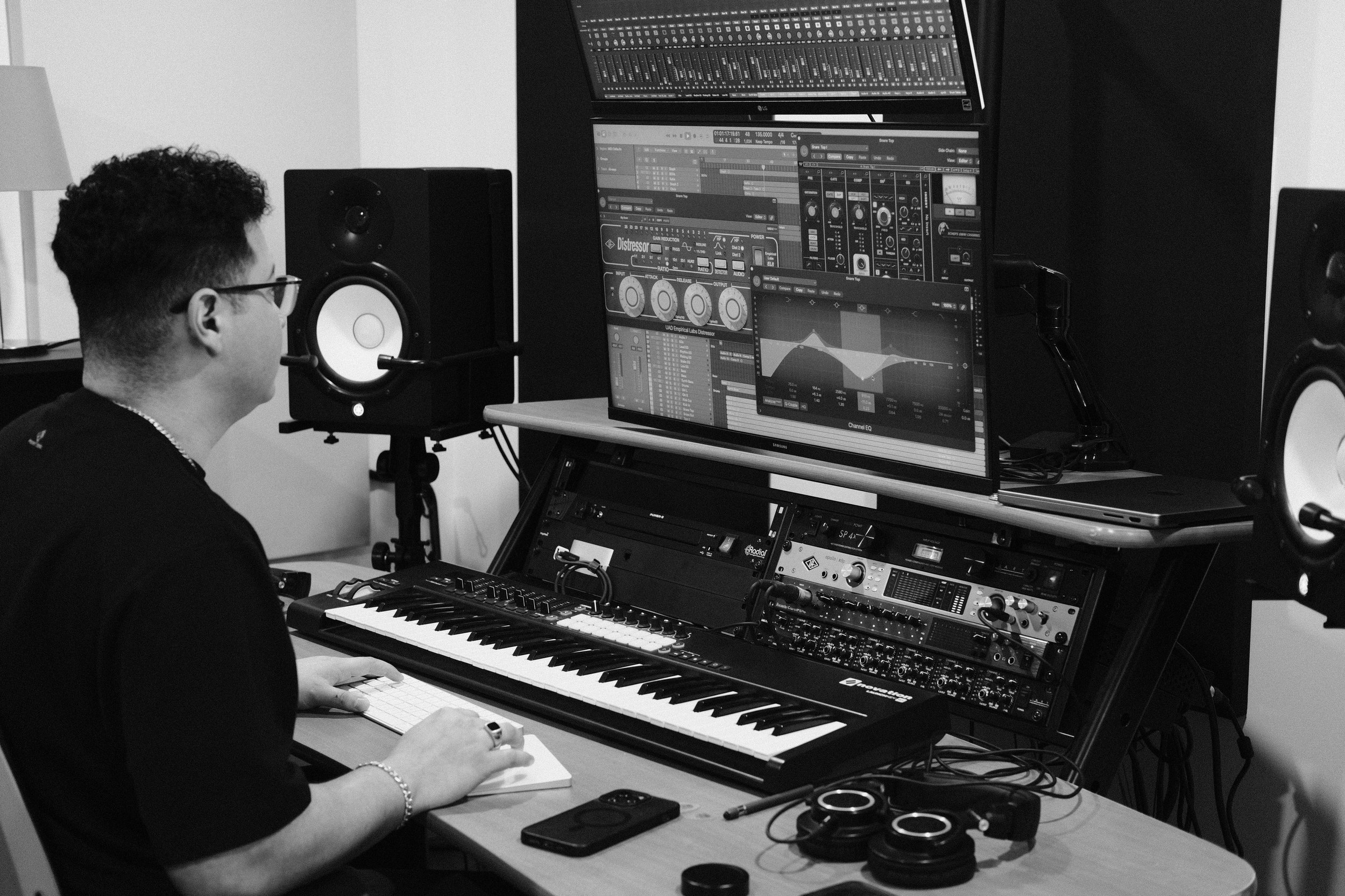 A person with glasses and curly hair sitting at a desk with a keyboard and a large monitor displaying music production software. Several studio speakers and audio equipment are visible in the room.