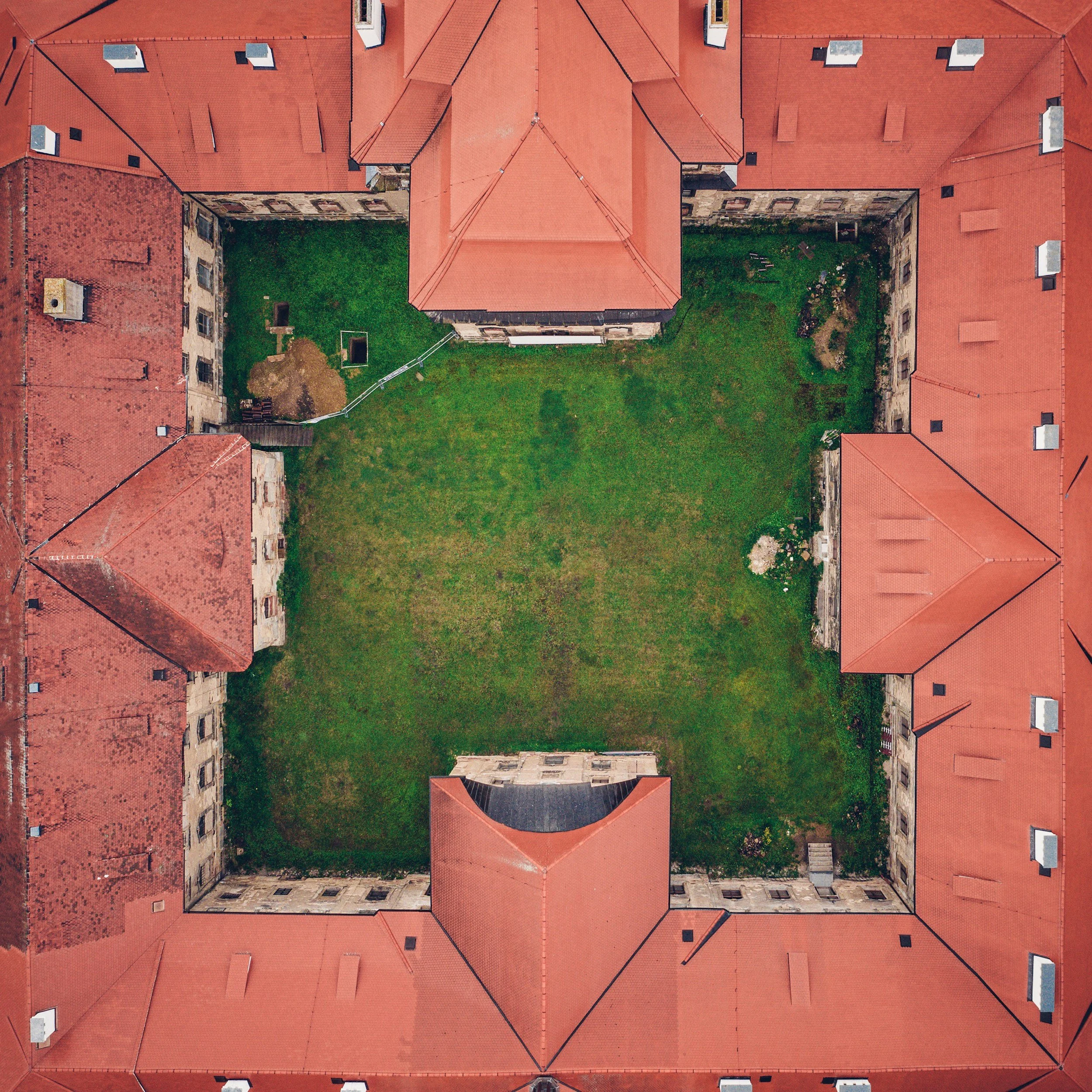 Aerial view of multiple red-tiled rooftops of buildings surrounding a grassy courtyard with some plants and trees.