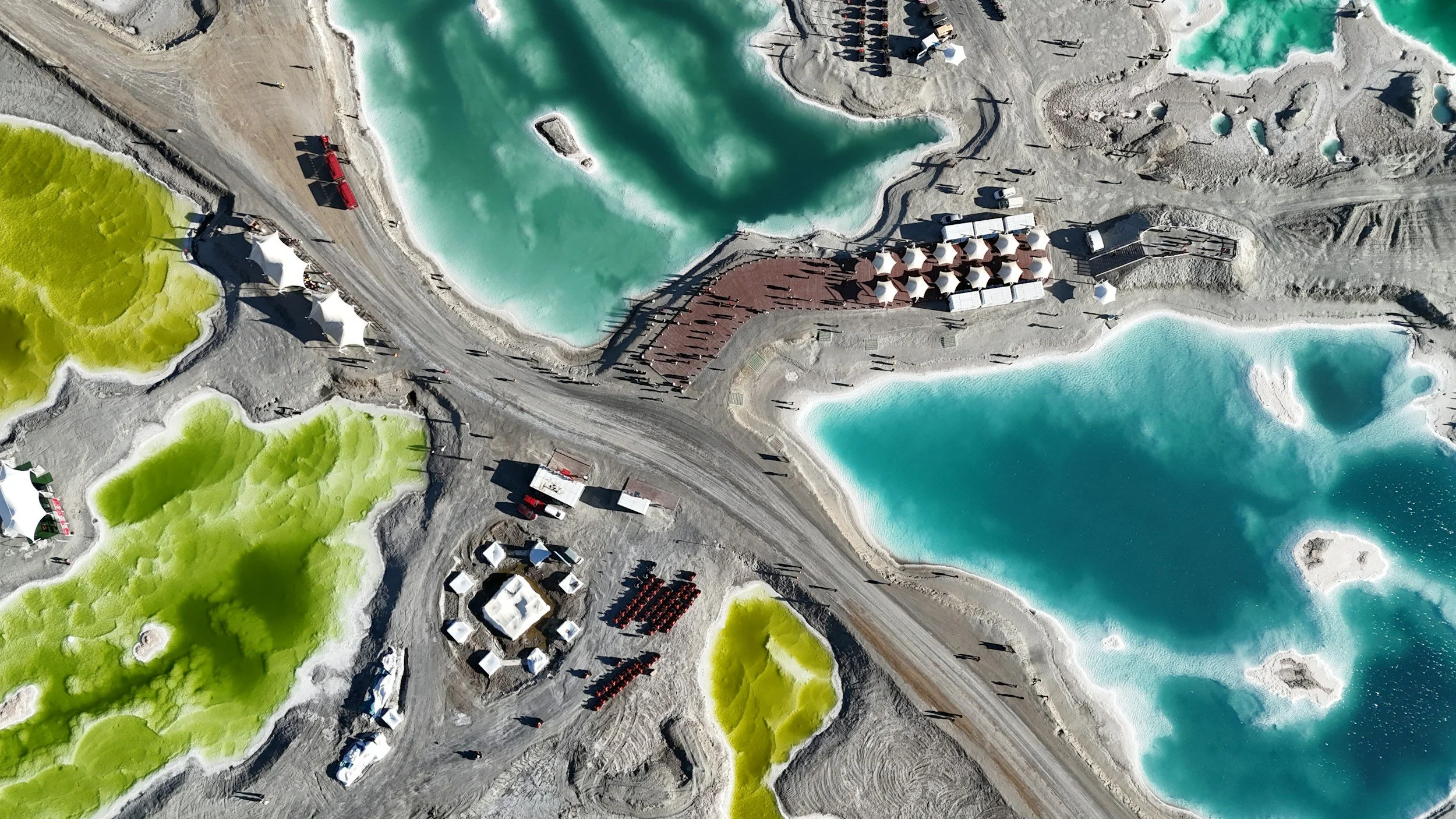Aerial view of a mining site with vibrant green and blue pools, tents, roads, and small structures, surrounded by barren landscape and scattered people.