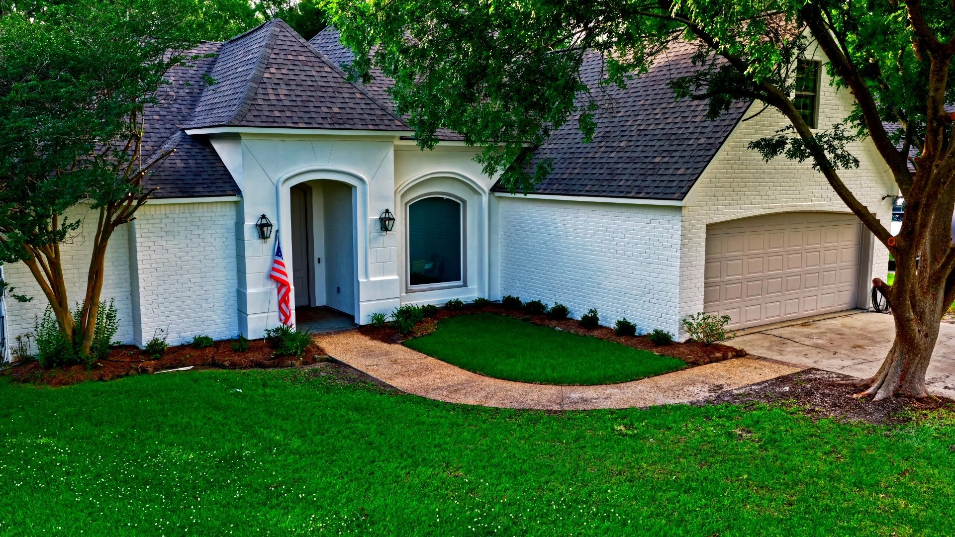 Front view of a white brick house with a brown garage door, a small porch, and a curved walkway surrounded by green lawn and trees.