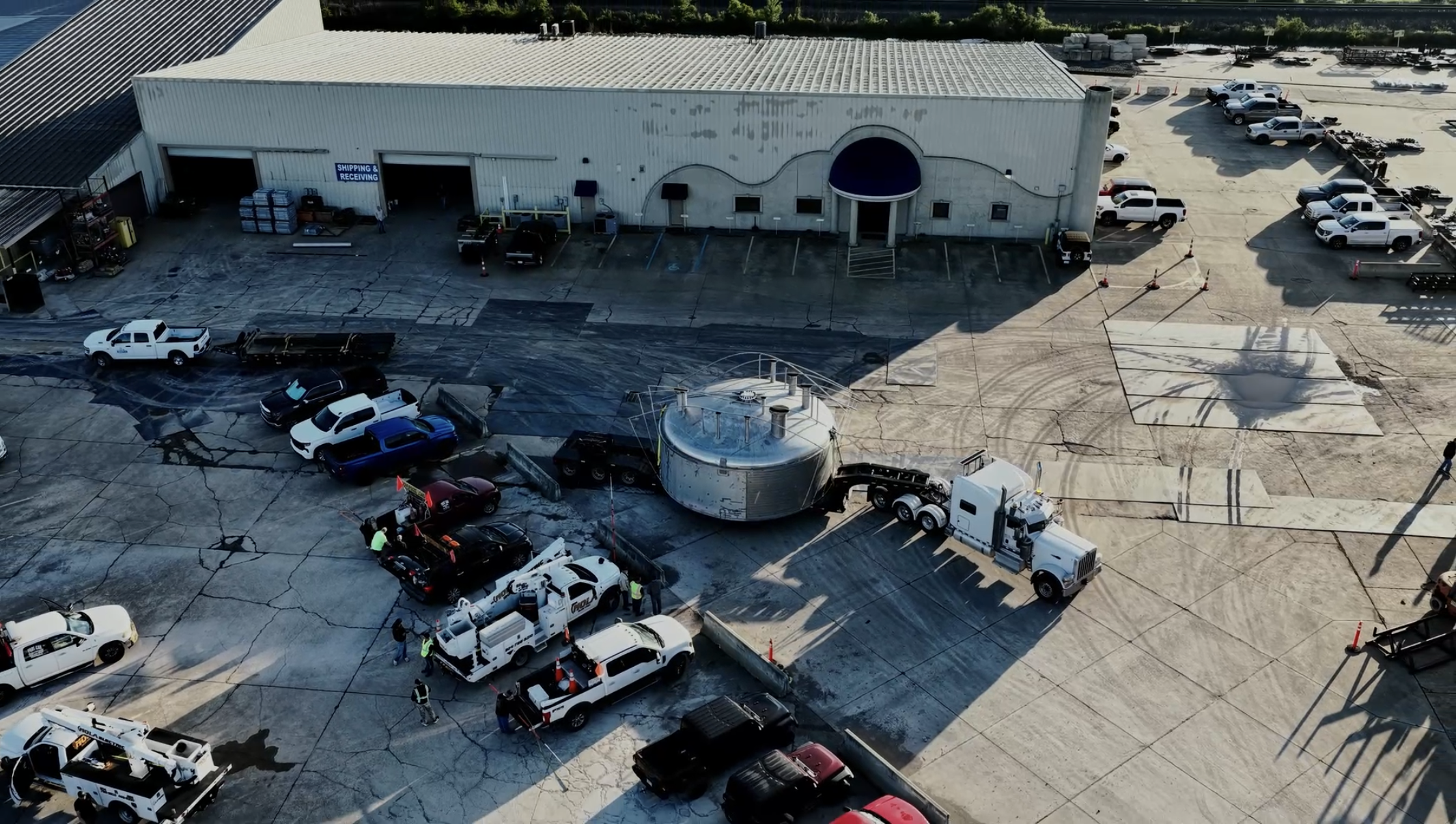 An aerial view of a large commercial building with a parking lot. Several trucks and vehicles are parked, and some workers are visible on the ground, possibly performing maintenance or construction work.