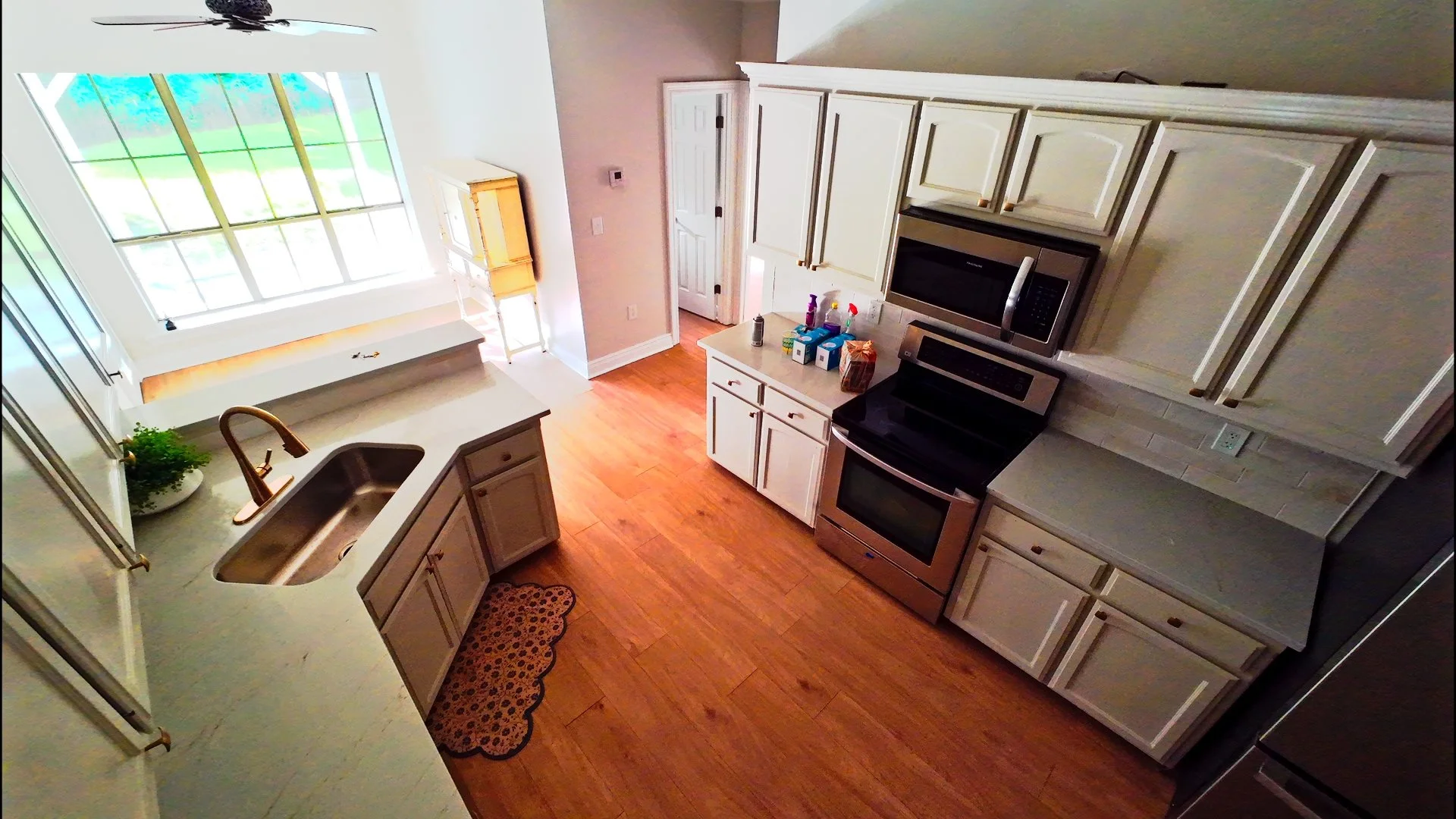A kitchen with white cabinets, stainless steel appliances including a microwave and oven, a white countertop with a sink, and a large window with a view of greenery outside. There are various items on the counter and a small plant by the sink.