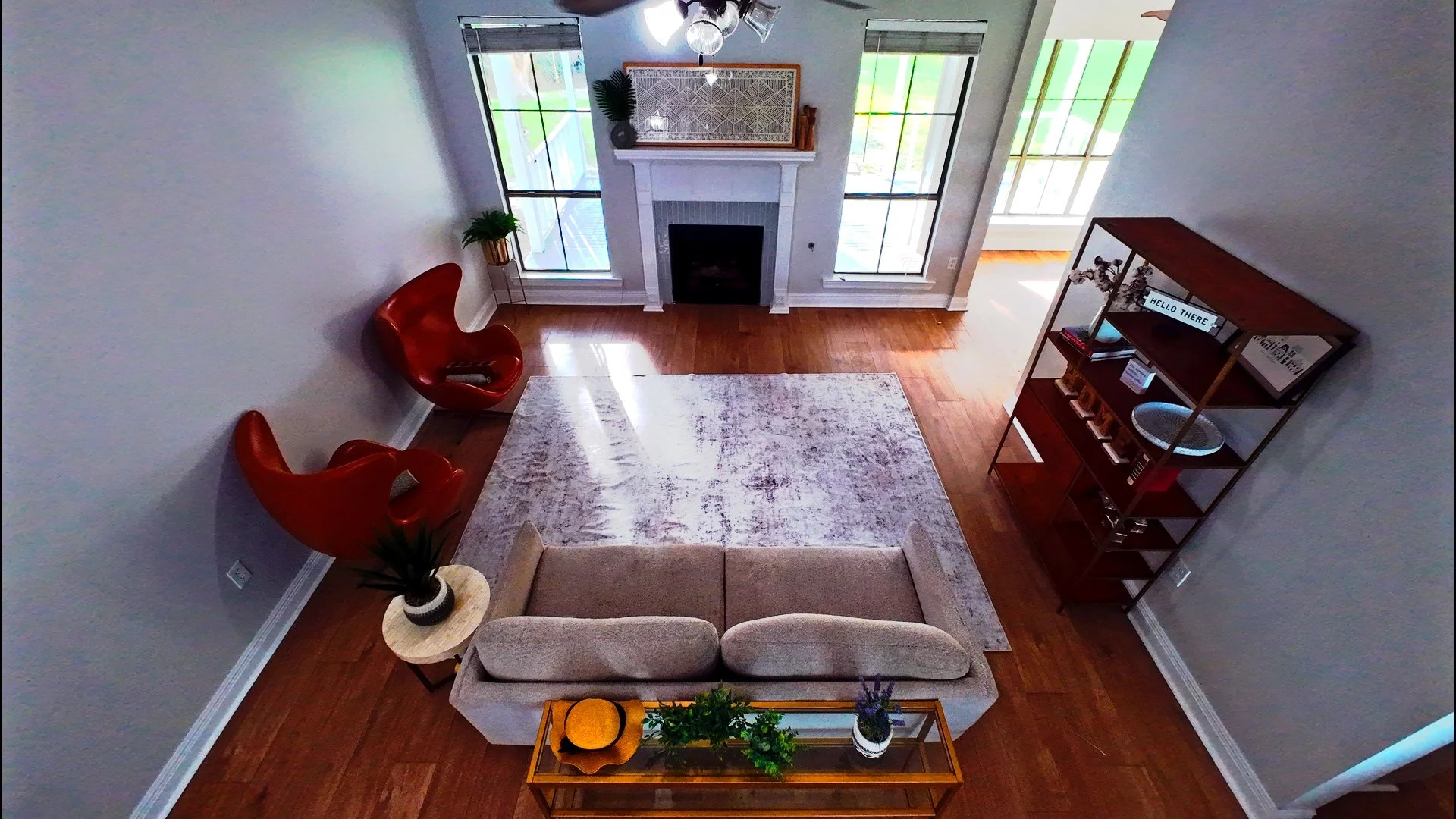Living room with a beige sofa, two red armchairs, a white area rug, a wood coffee table with decorative items, a white fireplace with art and plants above, large windows with blinds, and wooden flooring.