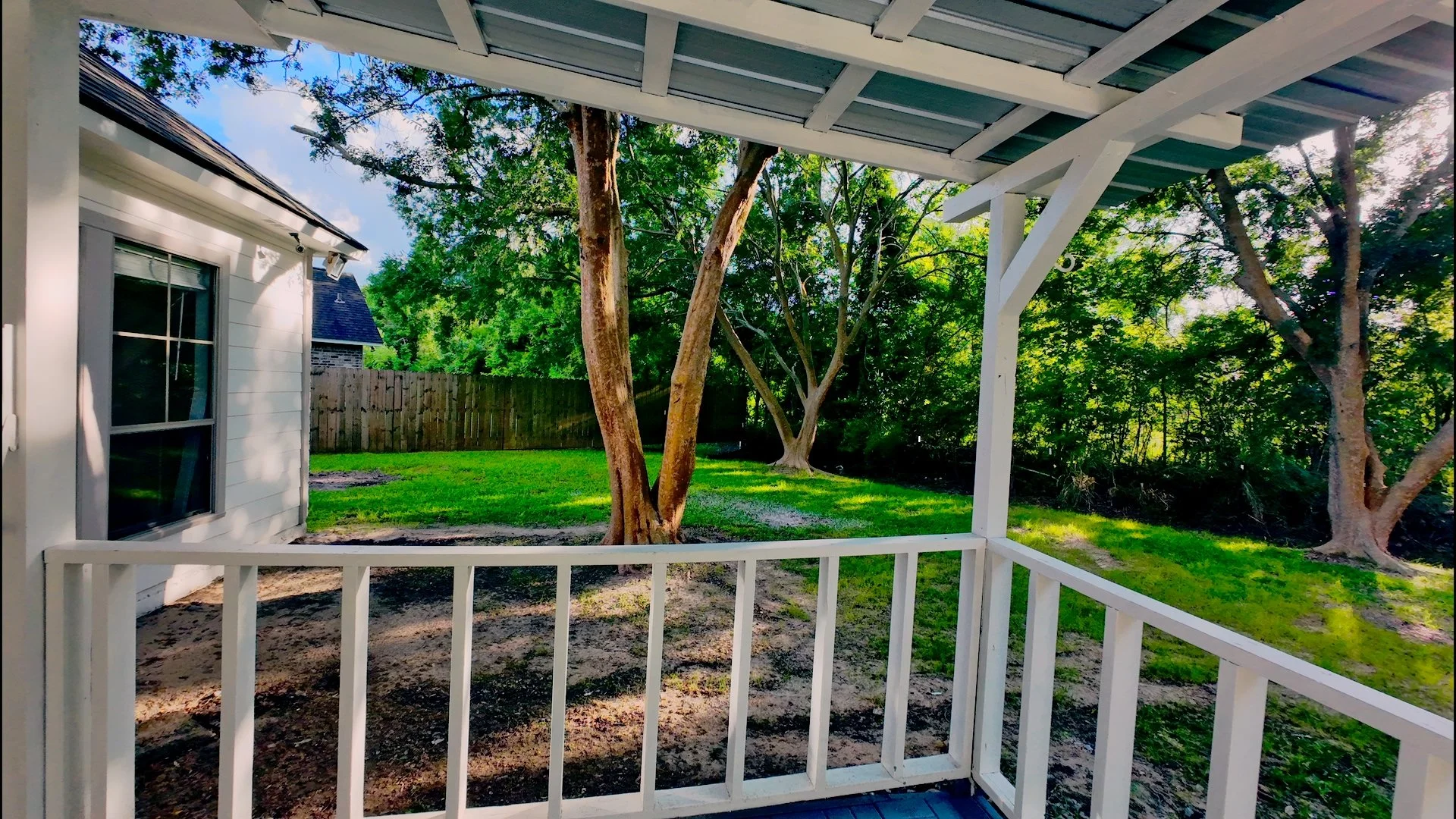 View from a porch showing a backyard with trees, a grassy lawn, a wooden fence, and neighboring houses.