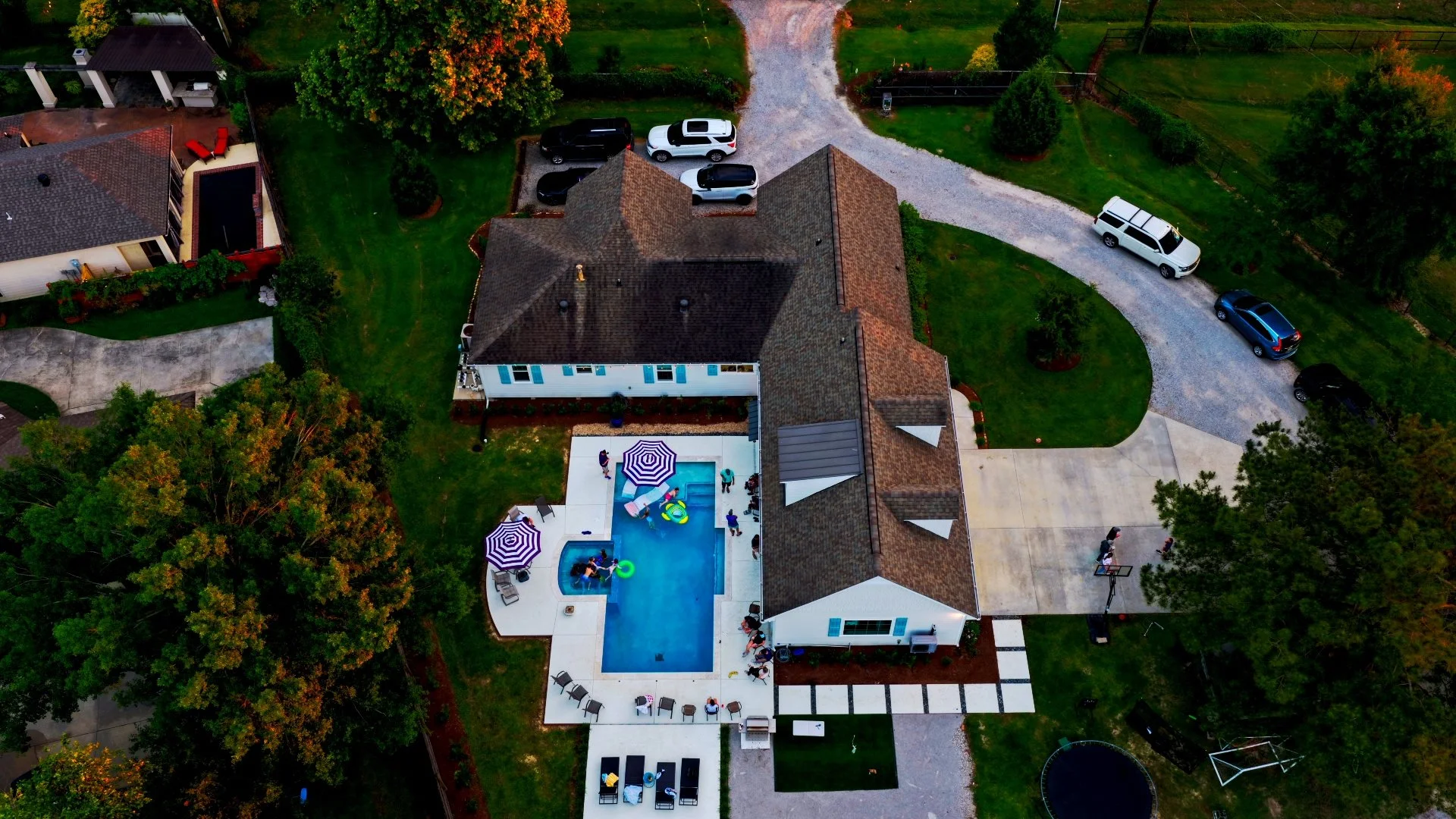 An aerial view of a suburban backyard featuring a swimming pool with umbrellas and people, a house with a gray roof, a driveway with parked cars, and surrounding trees and lawns.