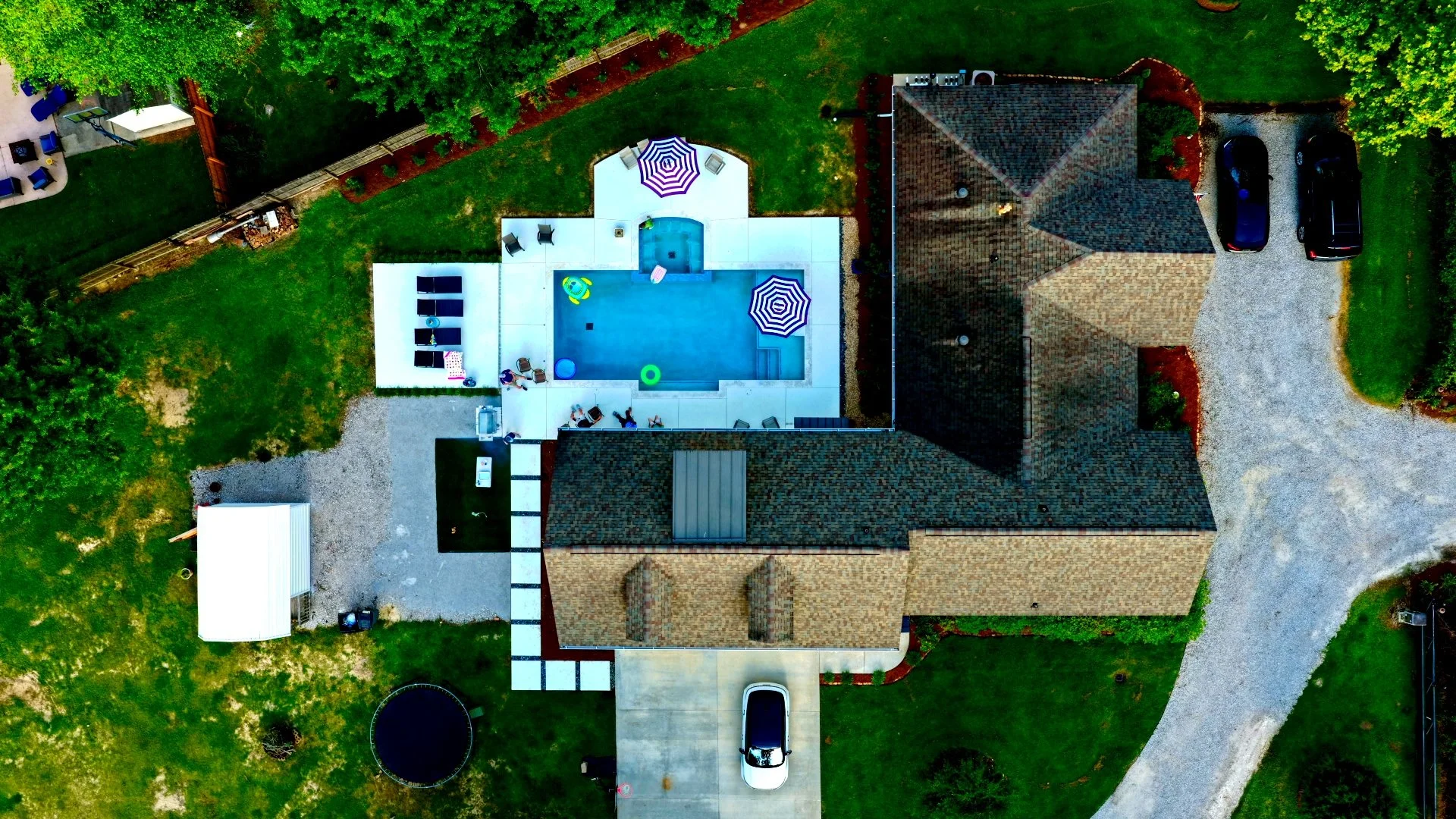 An aerial view of a house with a backyard featuring a pool with a striped umbrella, lounge chairs, and toys. The house has a driveway with two cars parked and a lawn with green grass and trees surrounding the property.