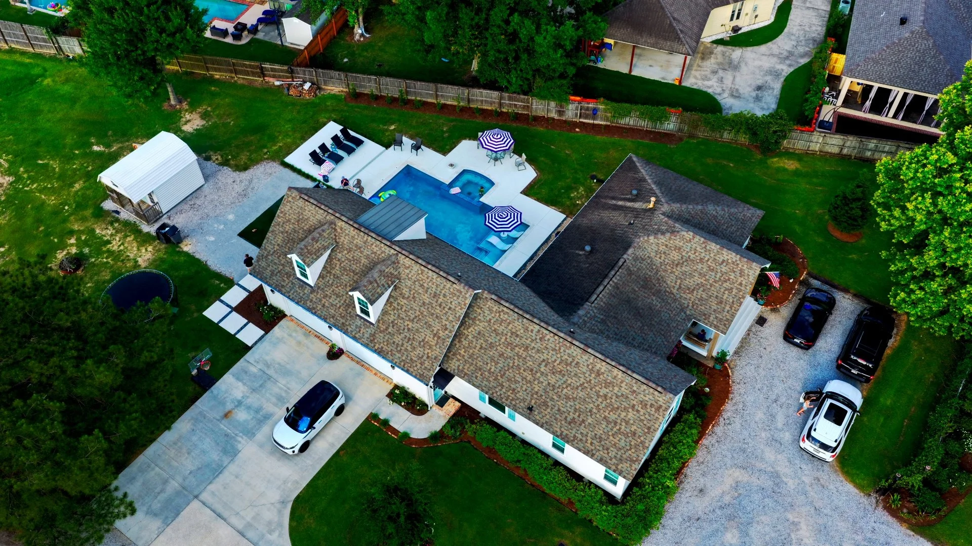 Aerial view of a suburban backyard with a swimming pool, patio umbrellas, and lounge chairs. There are several trees, a small white shed, a trampoline, and multiple parked cars. The house has a tiled roof and a driveway.