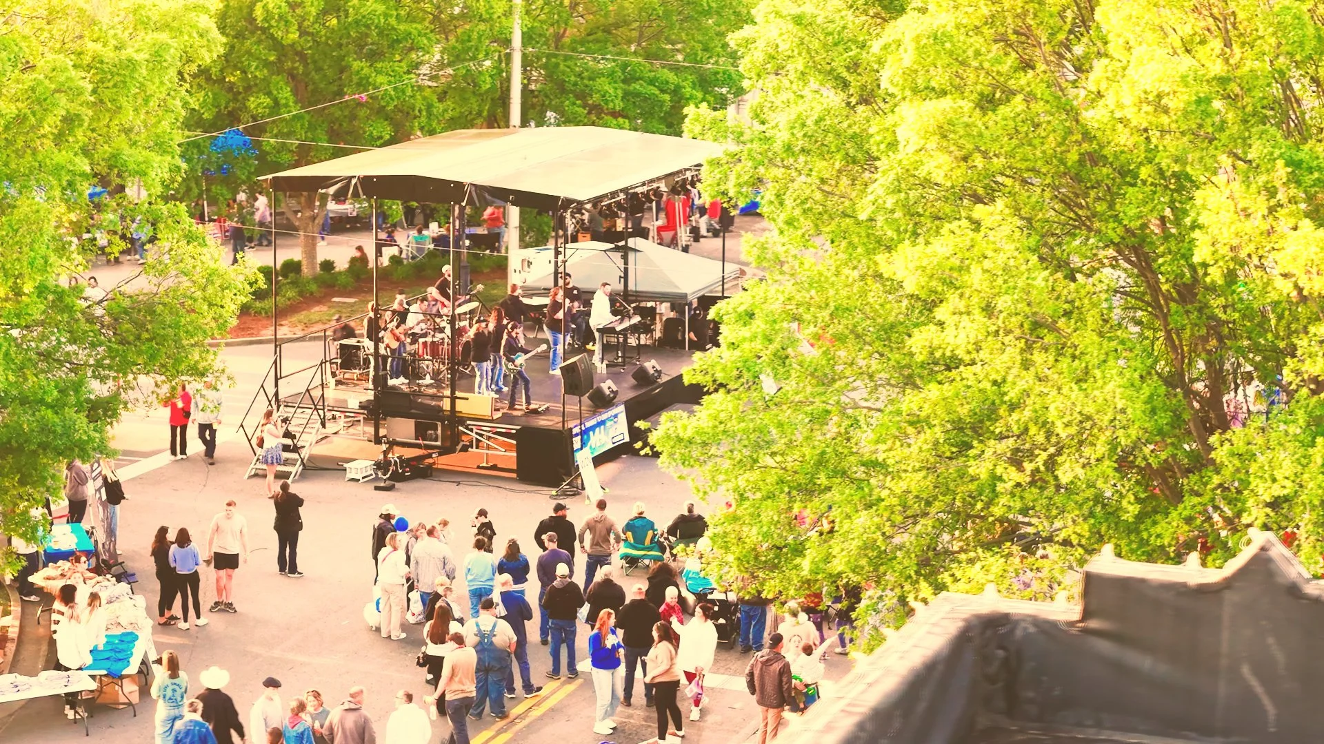 An outdoor music concert in a park with a band performing on a stage surrounded by large green trees, and people watching from the crowd.