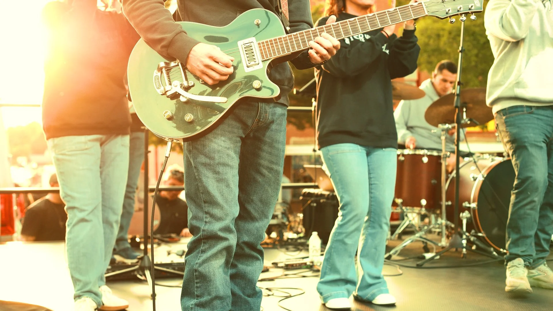 A group of musicians performing on stage outdoors in the sunlight, featuring a person playing a green electric guitar.