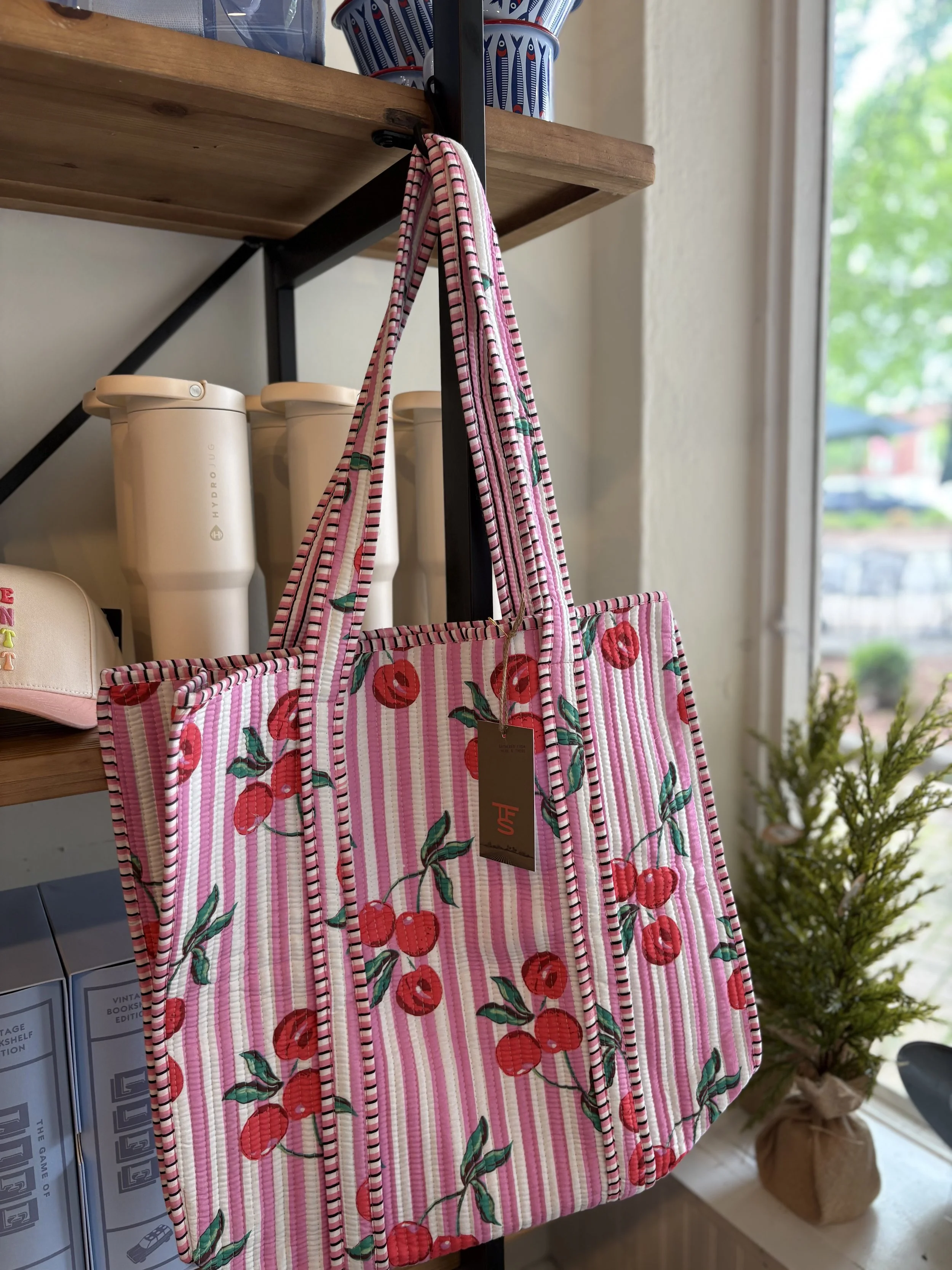 Pink and white striped tote bag with cherry print hanging on a black metal hook near a wooden shelf. In the background, there are white containers with green and red accents, and a window with a view of trees outside.