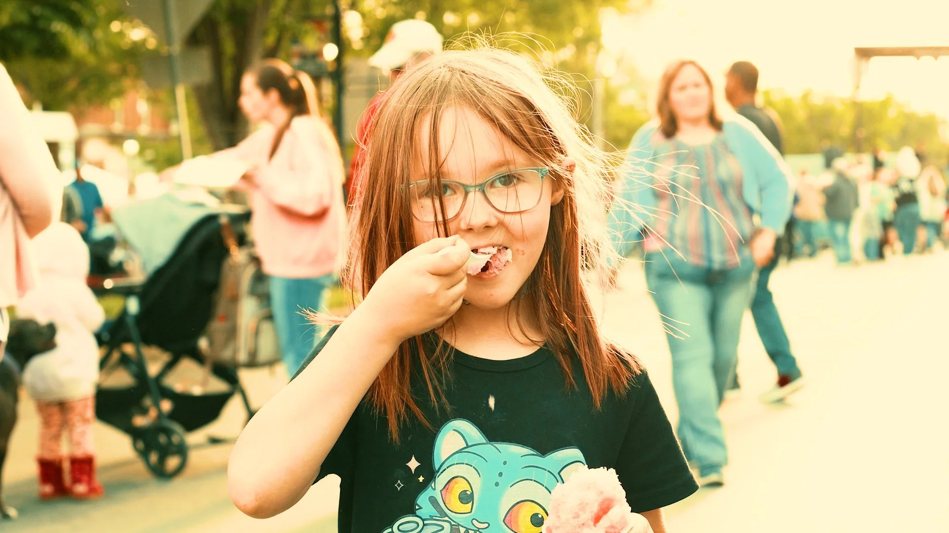 Young girl with glasses eating cotton candy at an outdoor event during sunset.