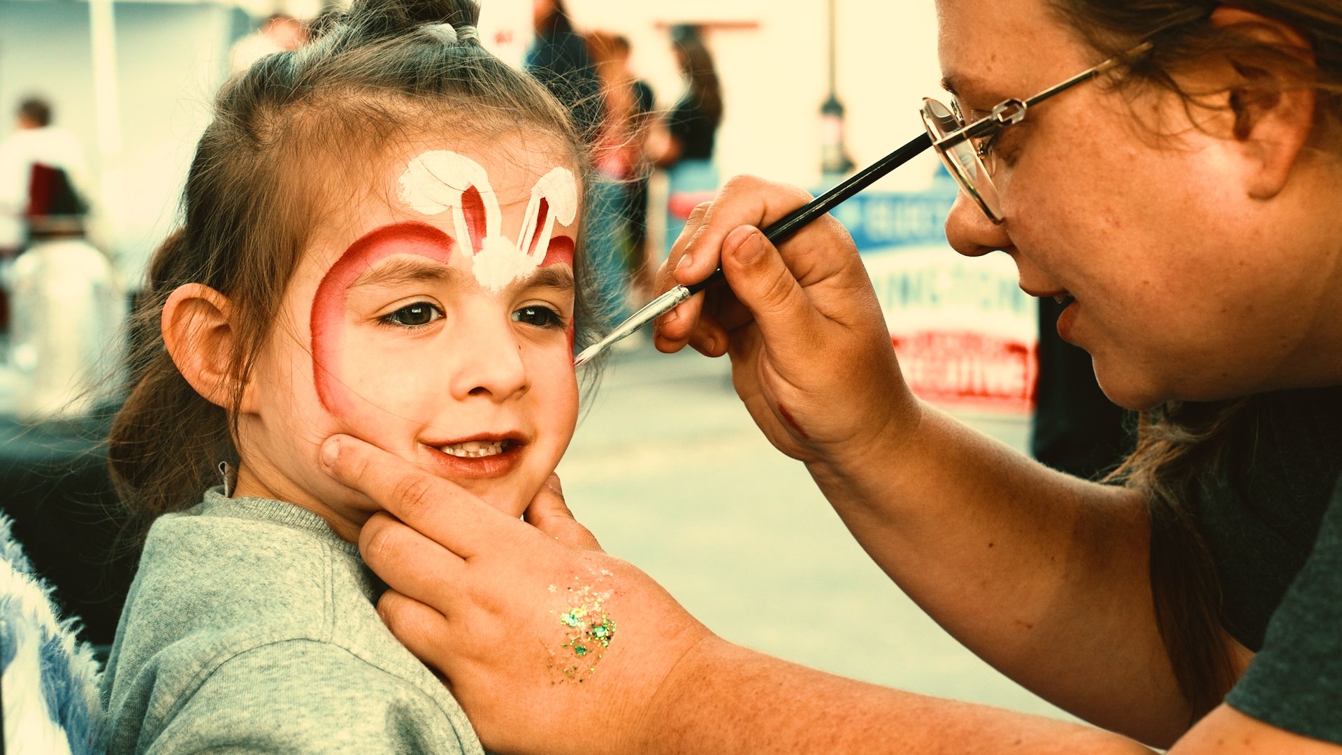 A young girl with face paint of a bunny gathers glitter on her face as an artist applies details with a paintbrush.